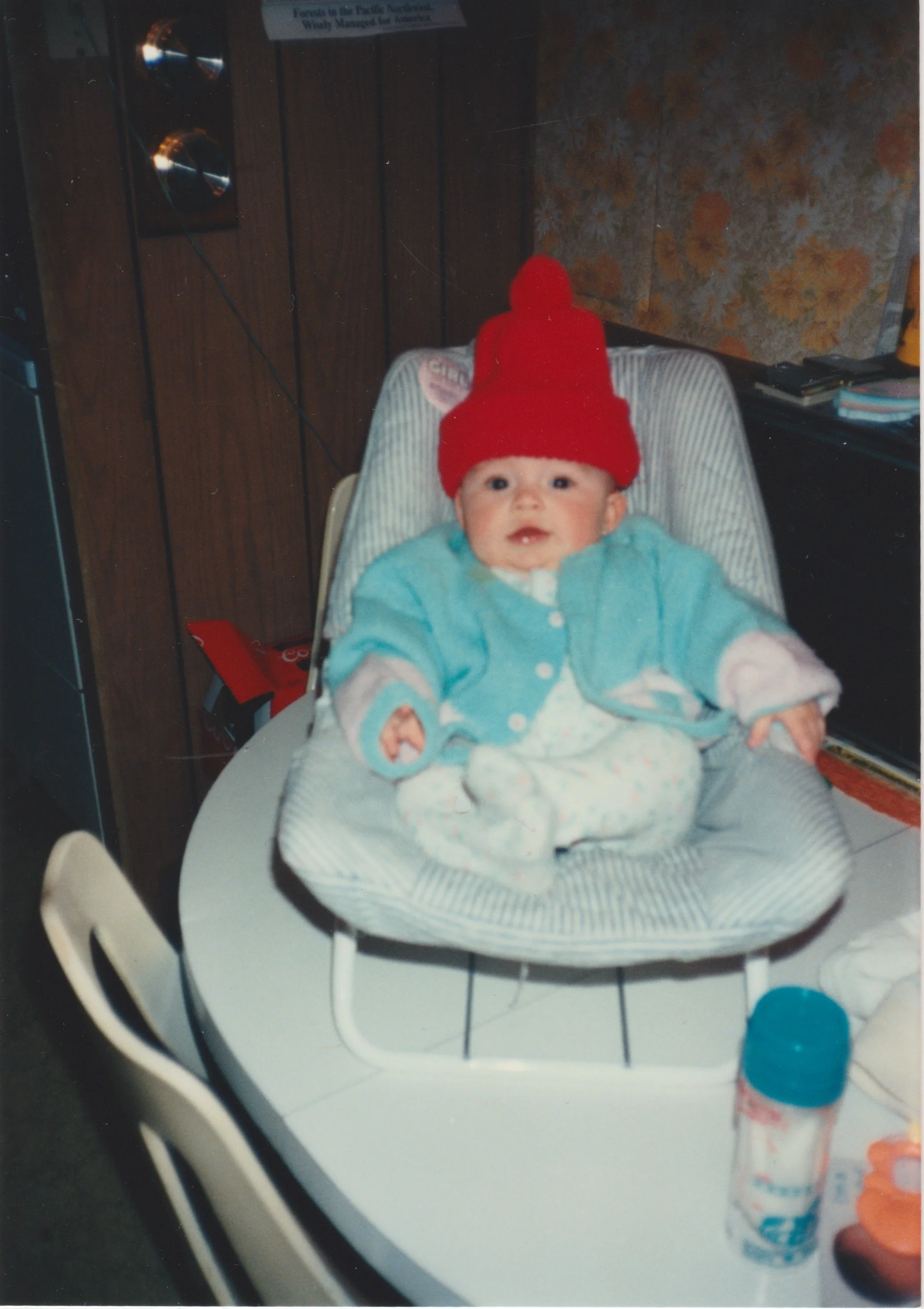 A baby sitting in a high chair on a dining table, wearing a red beanie hat, a light blue jacket, and white pajamas with small patterns. The background includes a wood-paneled wall by a stereo speaker, and the table has various items on it, including a blue-capped container and a small toy.
