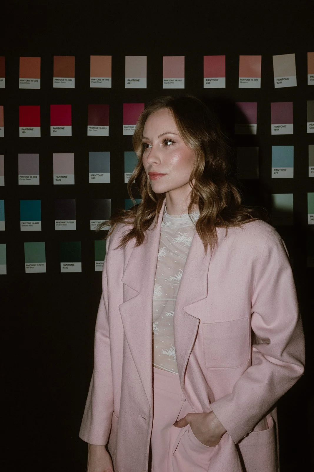 A woman with wavy brown hair and light makeup wearing a pink blazer and top stands in front of a wall displaying Pantone color swatches.
