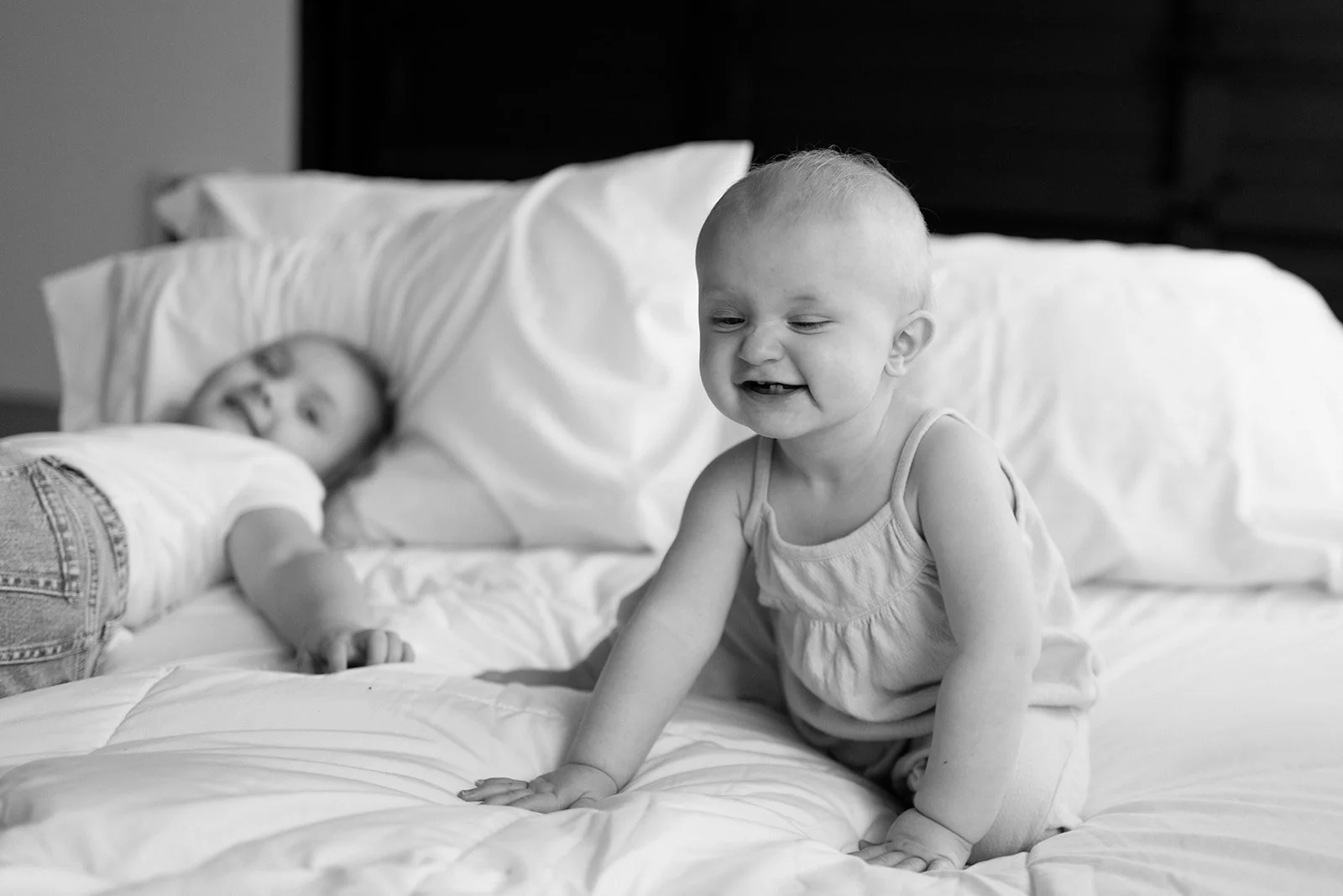 Two young children with bald heads, one sitting on the bed and smiling, the other lying down, both on a bed with white sheets and pillows in a bedroom.