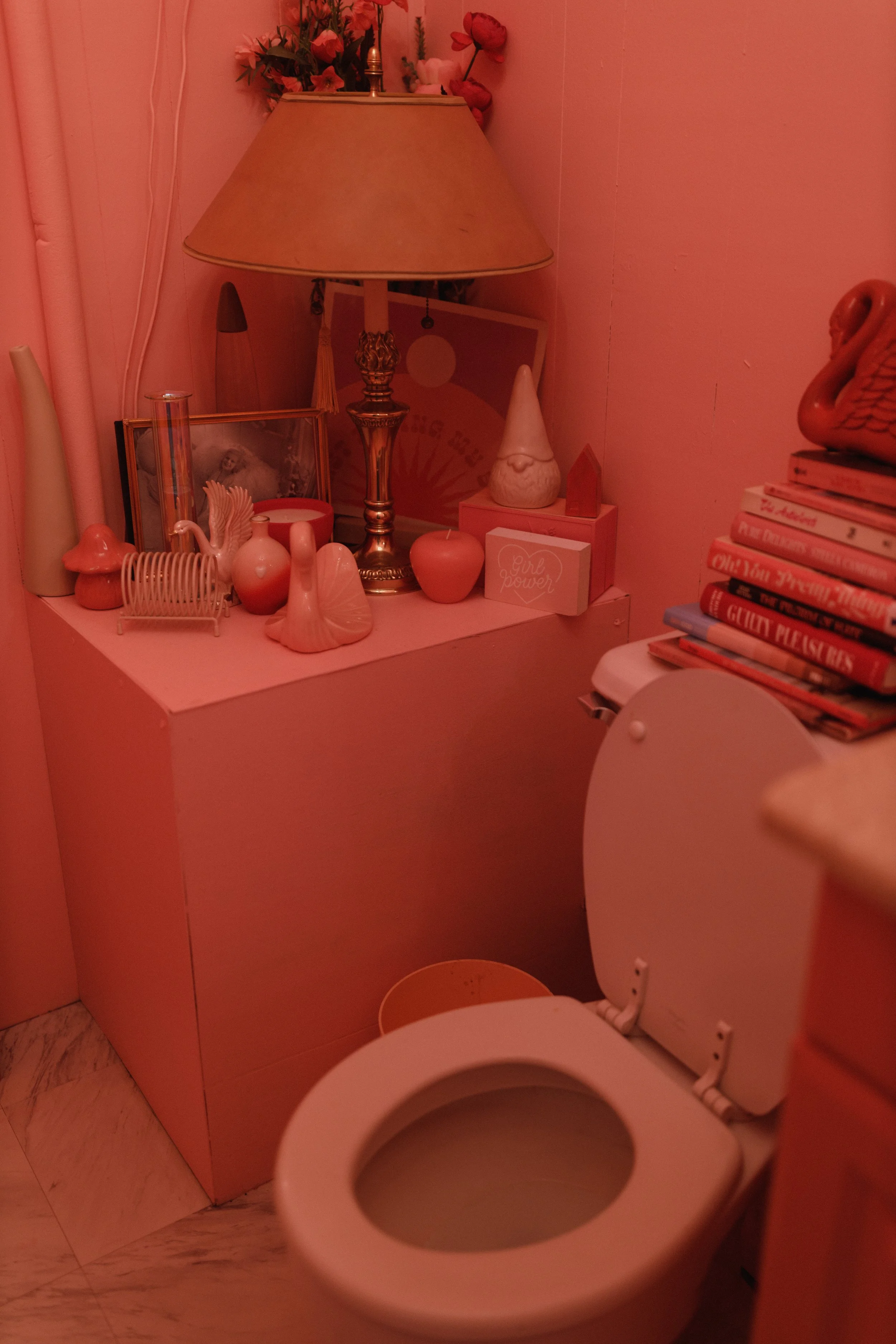 A bathroom with pink decor, including a pink vanity, pink lamp, and pink decorative items. There are candles, a framed picture, and various small decor pieces on the vanity. Books are stacked on a surface next to the toilet, which is open.