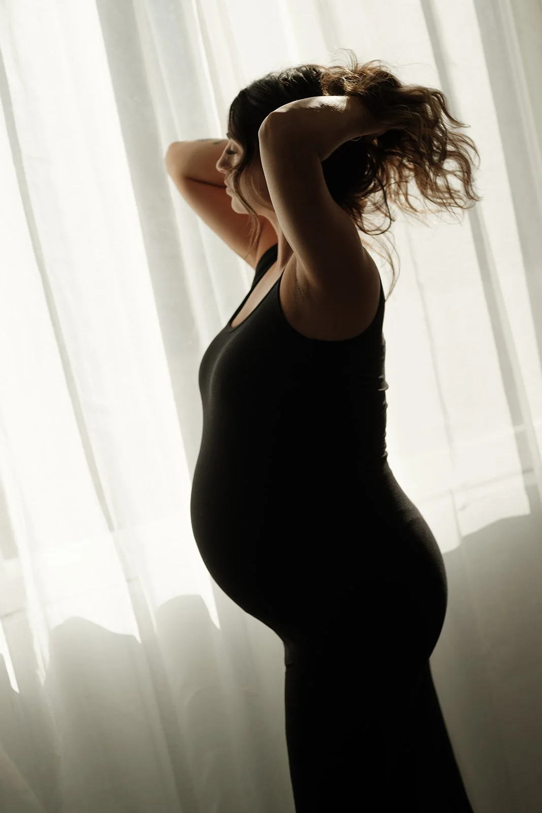 A woman with wavy hair in a black sleeveless dress, standing near white curtains, with sunlight creating a silhouette effect.