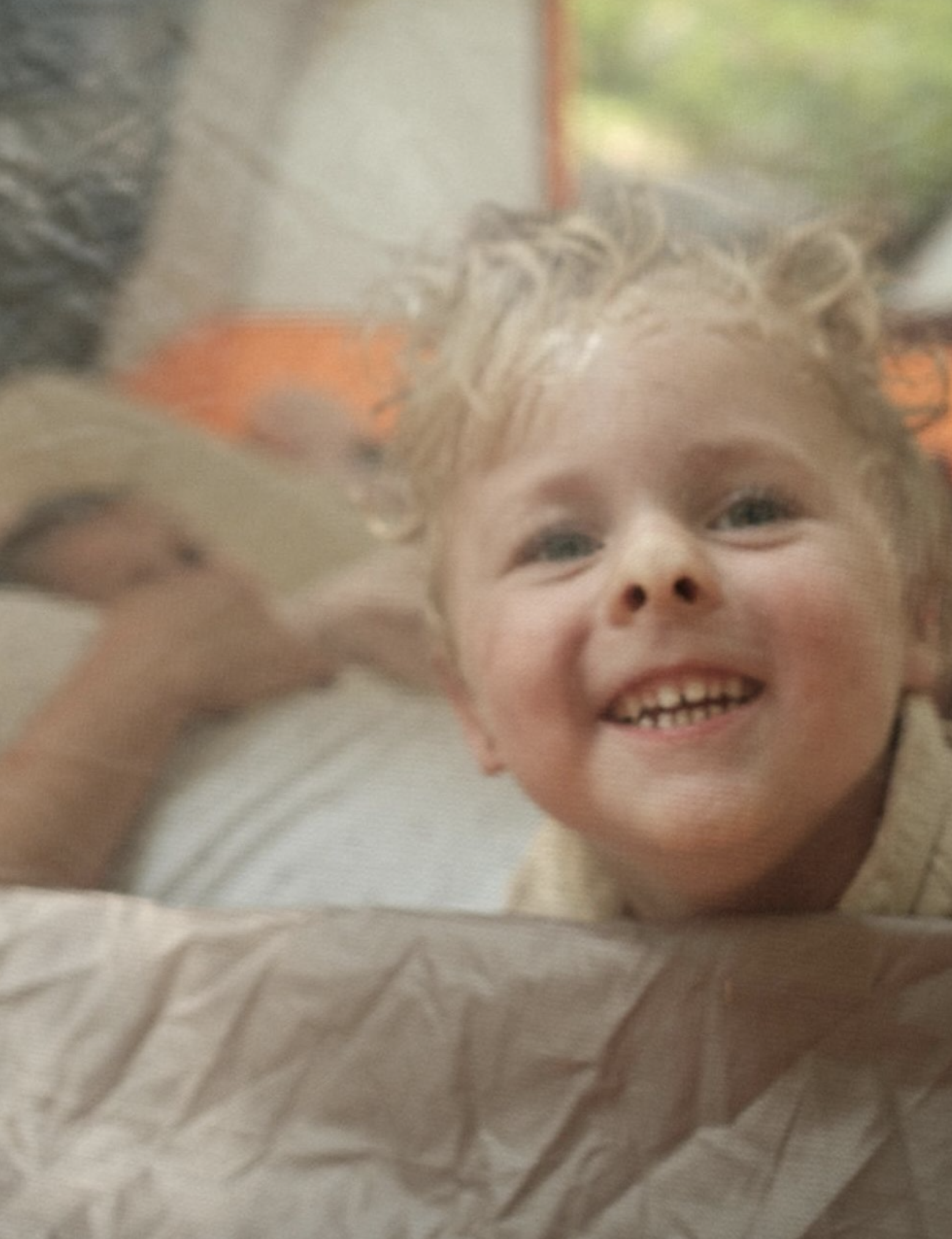 Close-up of a young child with curly blonde hair, smiling widely, with a person in the background.