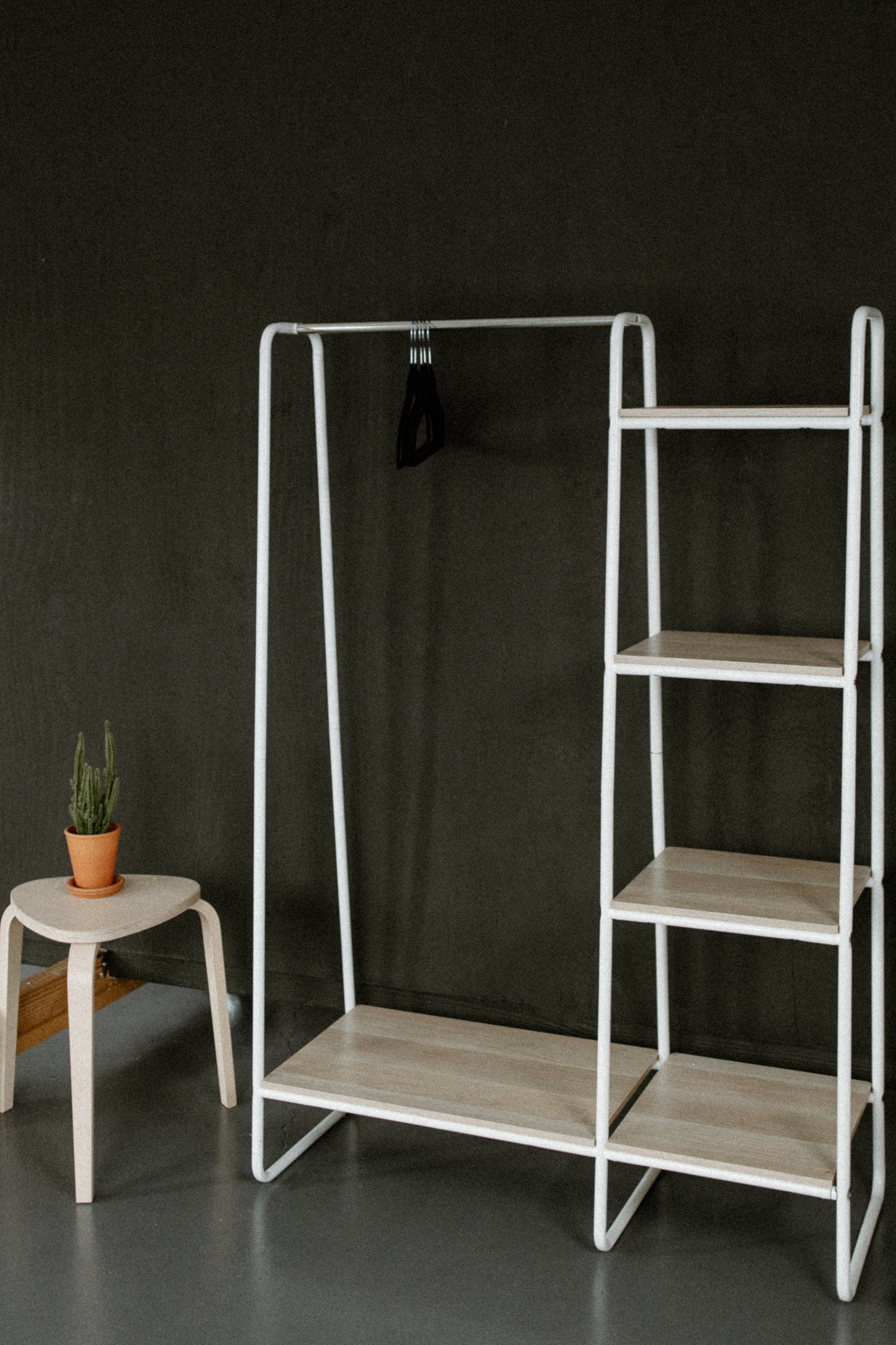 A minimalist room with a black wall and a gray floor. There is a white clothing rack with a black hanger hanging on it, and a tall, narrow shelving unit attached to the rack. To the left, there is a small white stool with a small potted cactus plant 