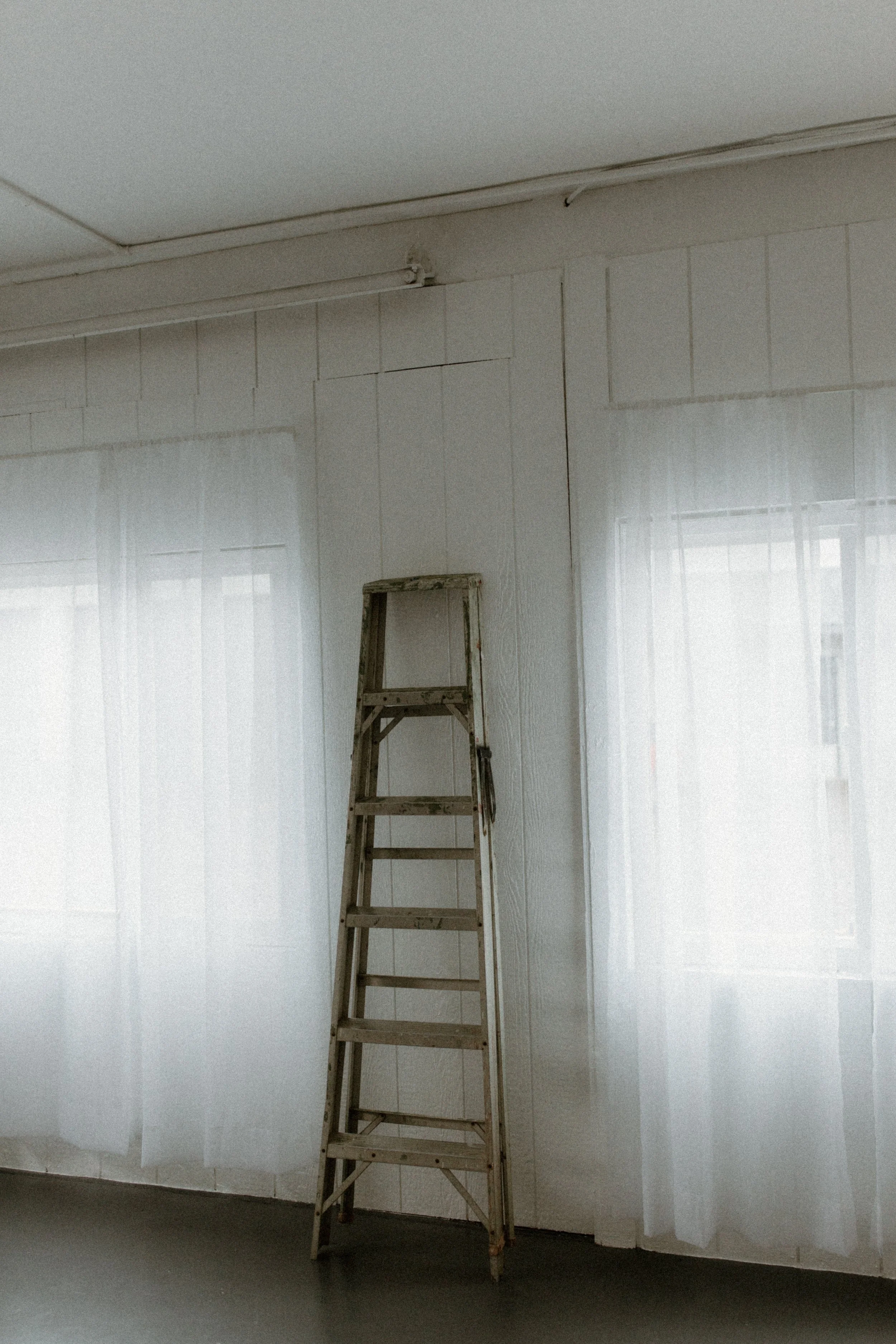 A wooden step ladder standing in a room with white walls and sheer curtains covering windows.