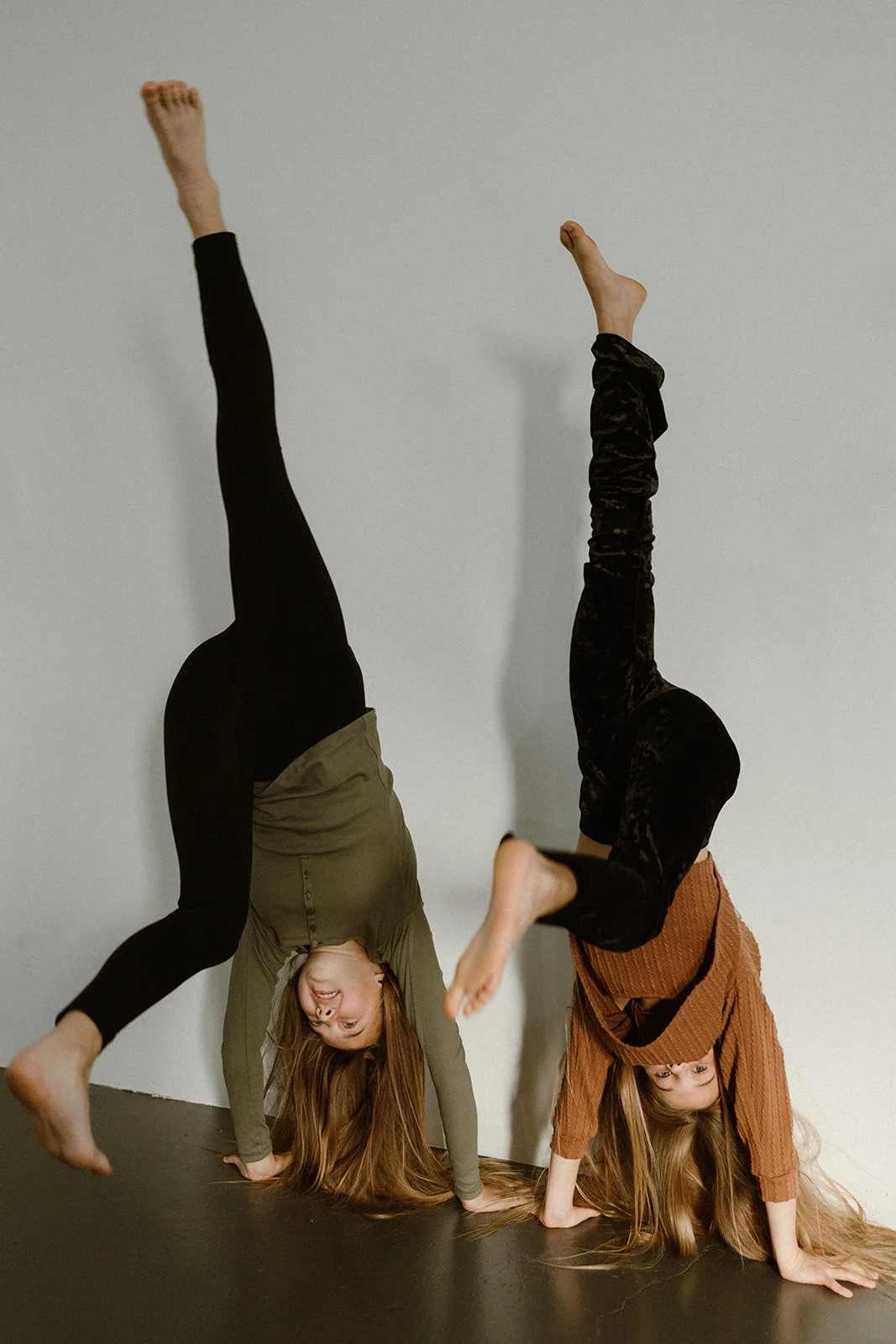 Two women performing handstands against a white wall, one wearing a green top and black leggings, the other in a brown sweater and black pants, with their hair hanging down.