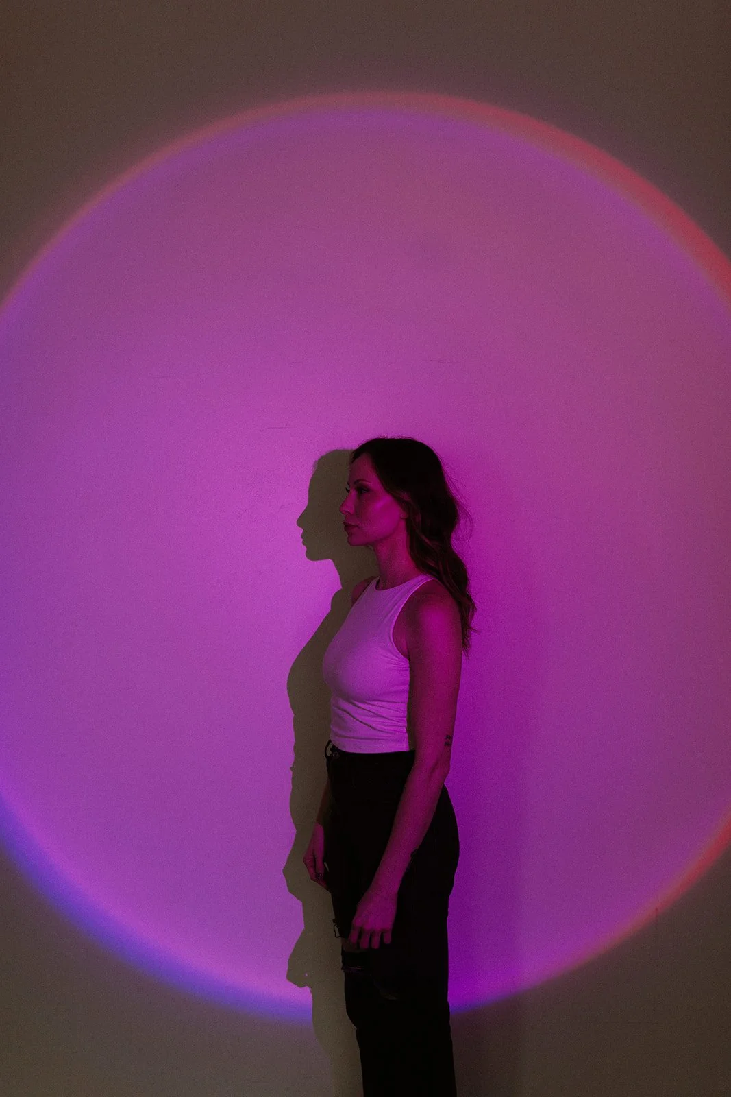 A woman standing against a wall with a large purple and pink circular light projection behind her, casting a shadow on the wall.