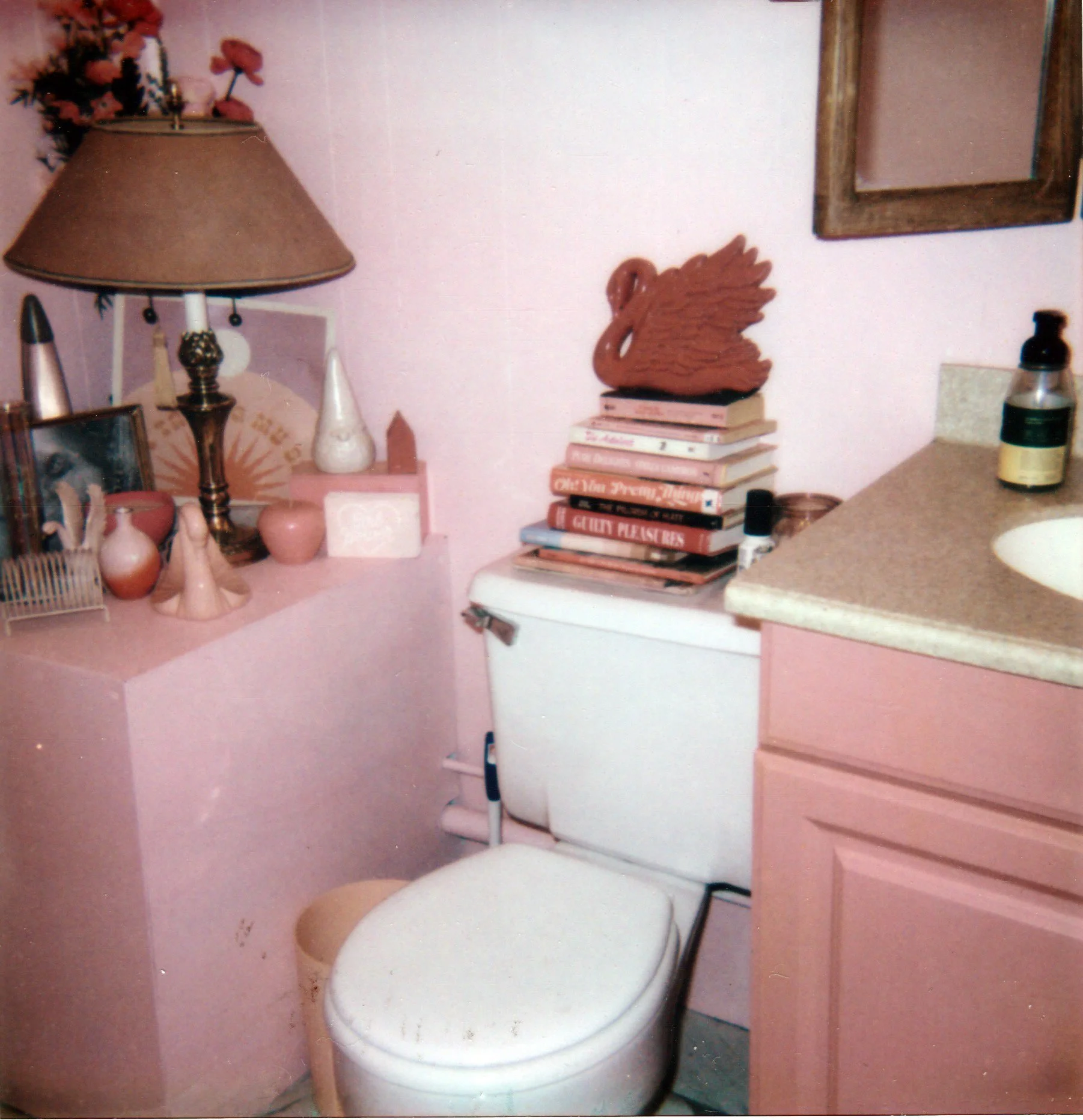 A small bathroom with pink walls, a white toilet, and a pink vanity with a beige countertop. The top of the toilet lid shows a stack of books with a wooden swan sculpture on top. Next to the toilet is a lamp with a beige shade and decorative pink vas