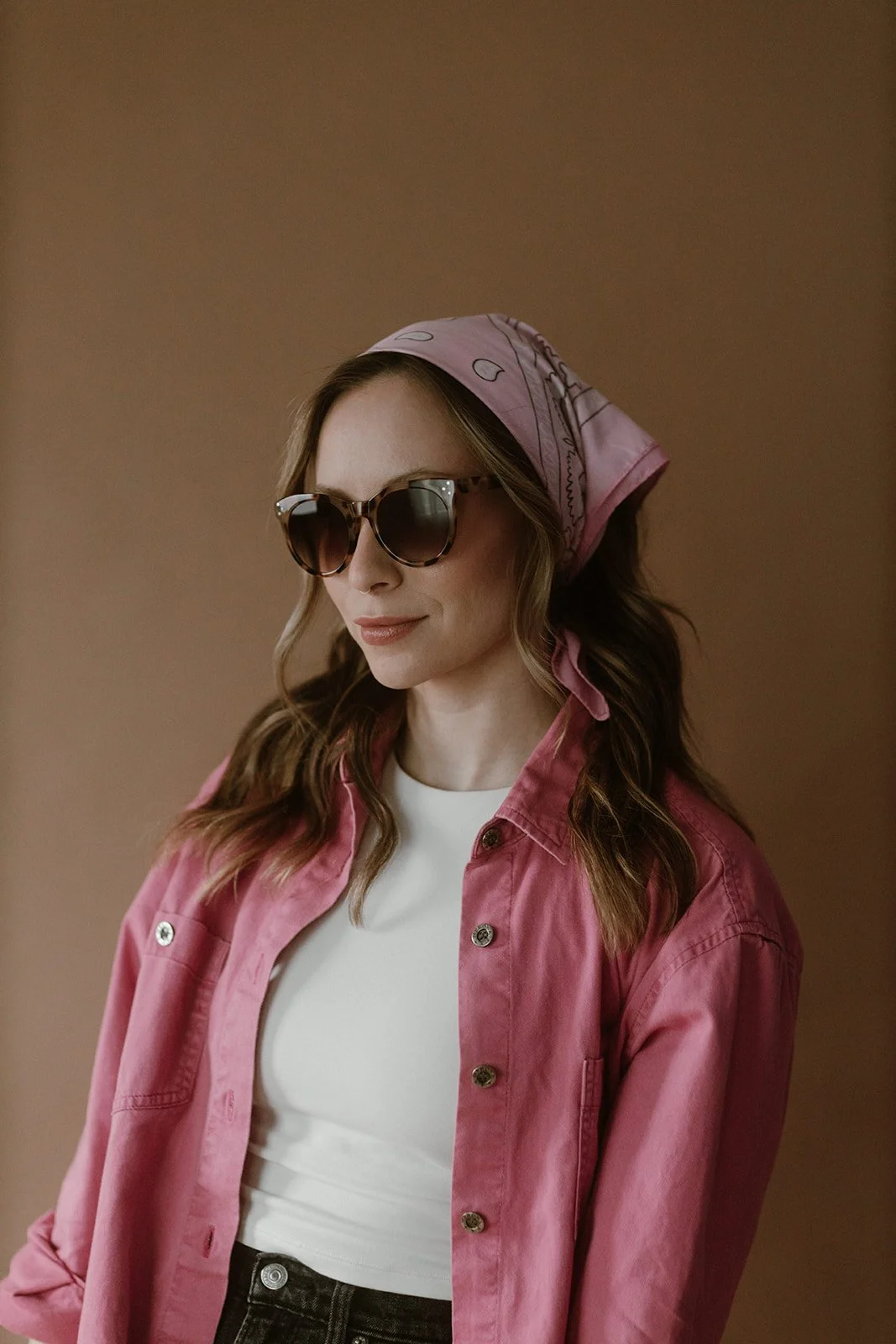 A woman with wavy brown hair wearing large sunglasses, a pink bandana, a pink denim jacket, and a white top, posing against a brown background.