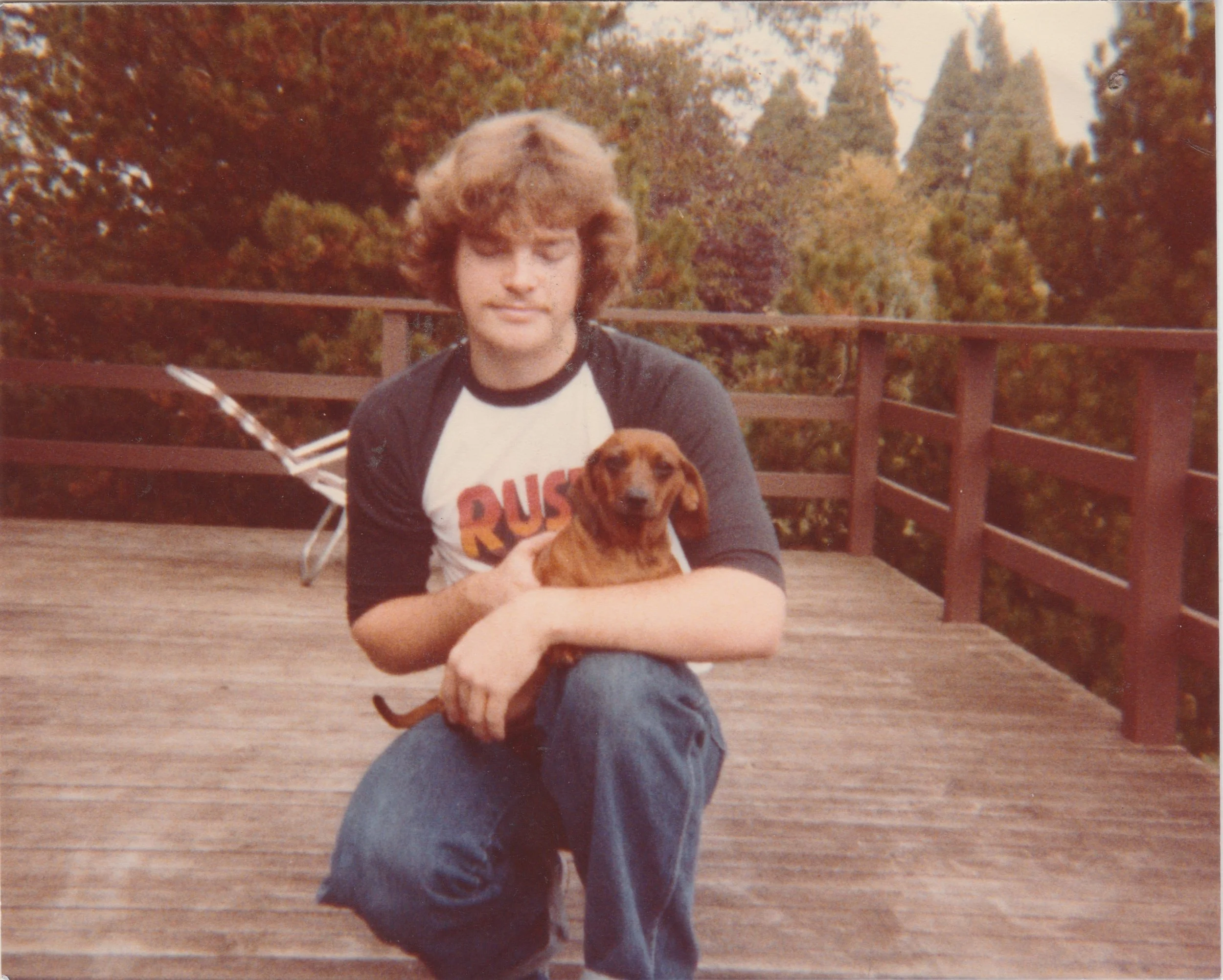 A young man with brown, wavy hair wearing a Rush band T-shirt and jeans, kneeling on a wooden deck, holding a small brown dachshund puppy. The background features a wooden railing and autumn-colored trees.