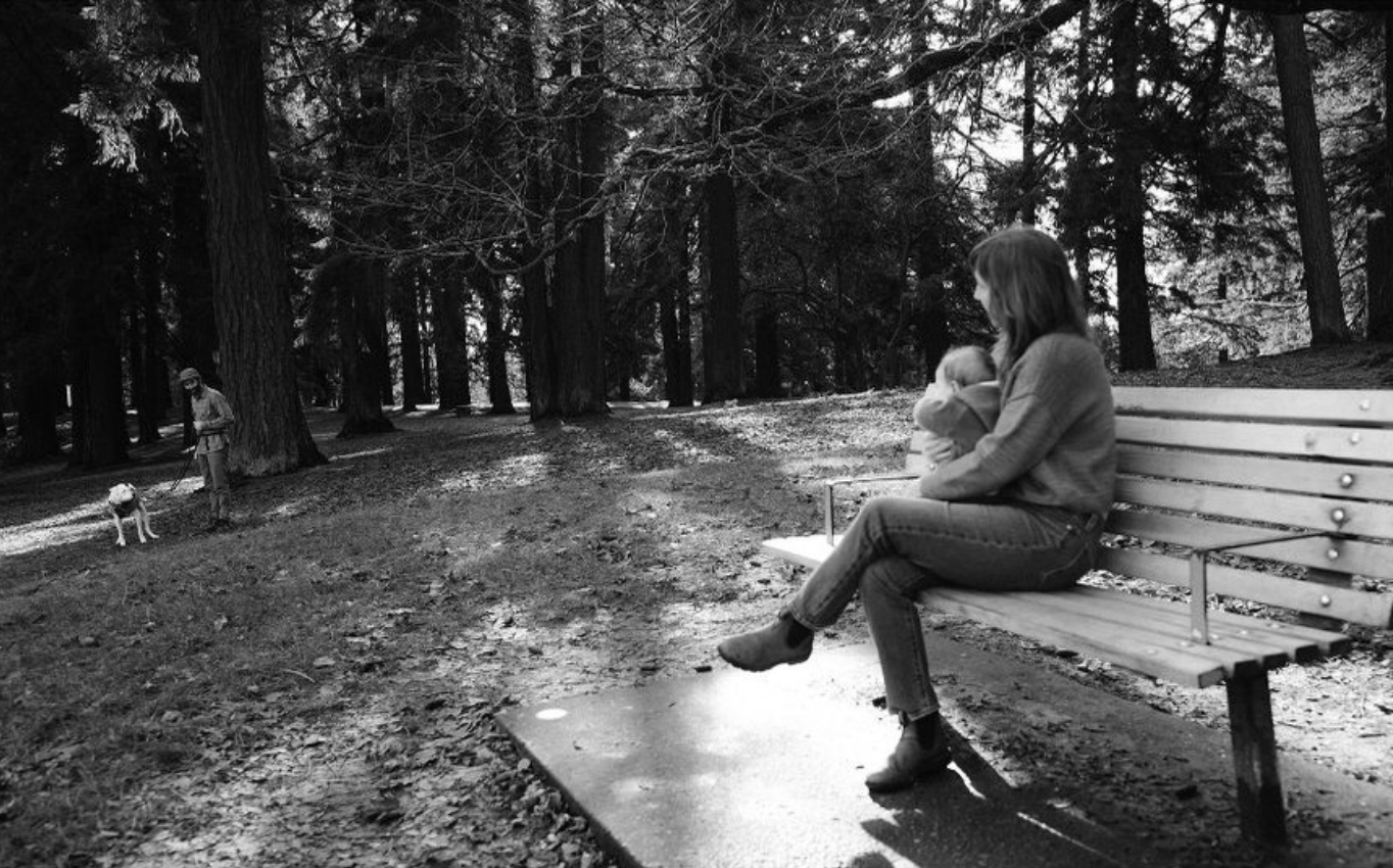 A woman sitting on a park bench holding an infant, with a man walking his dog in a wooded park area.