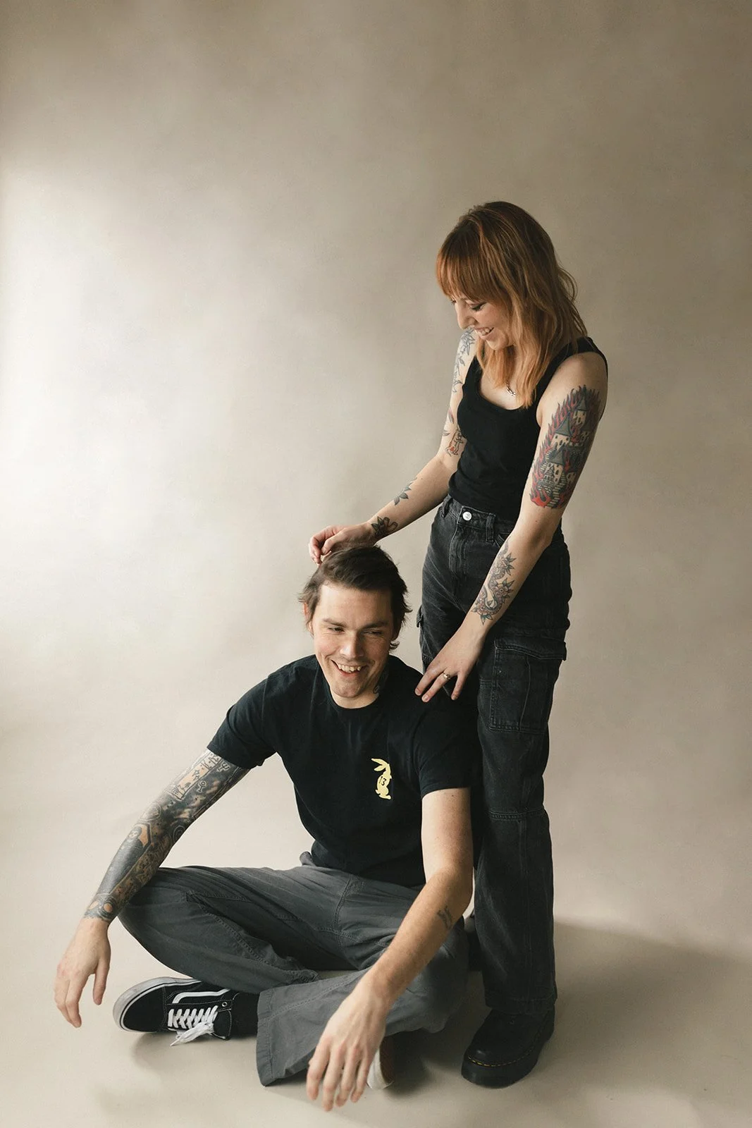 A woman standing behind a crouching man, smiling and gently petting his hair, both with tattoos, against a plain light-colored background.