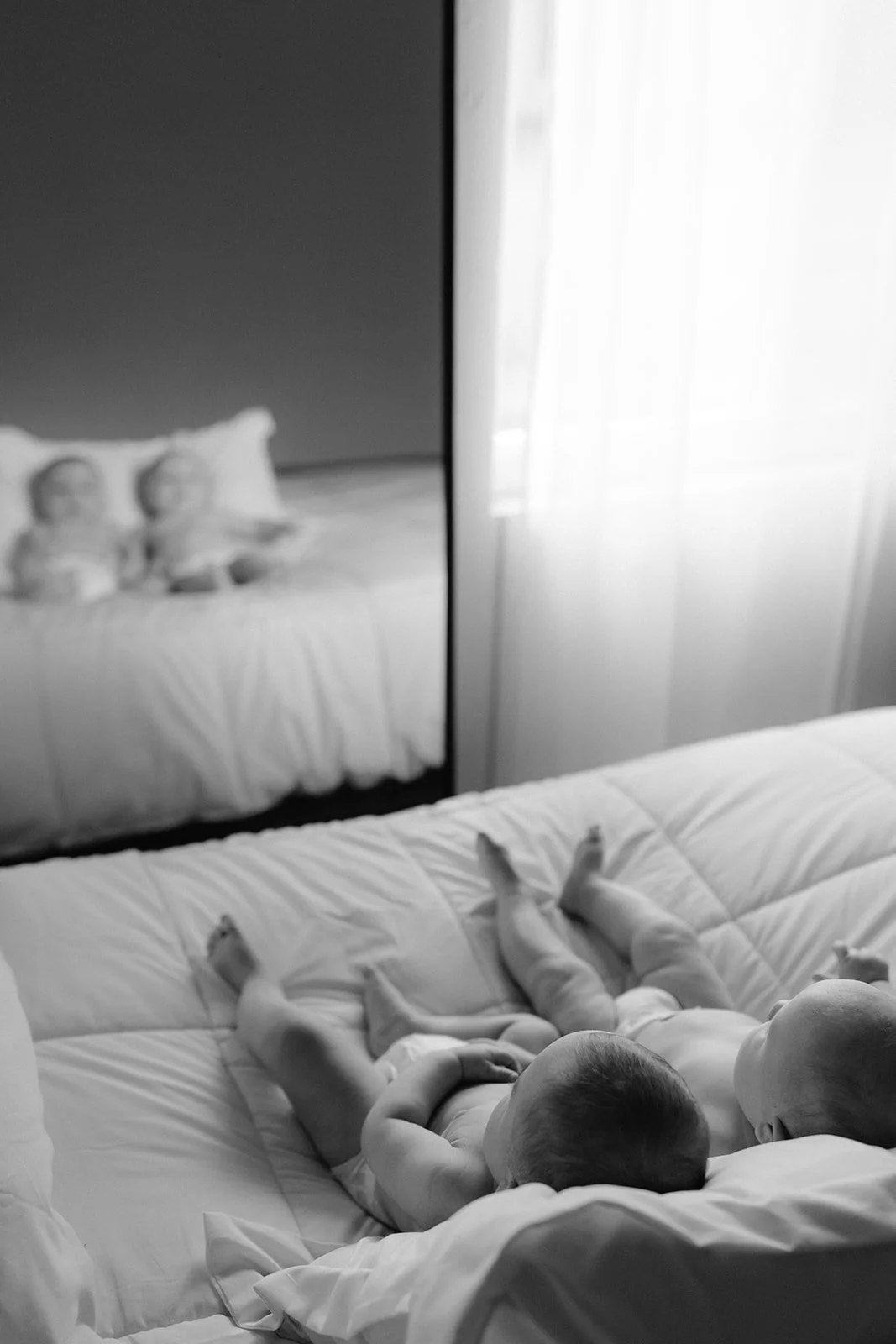 A black-and-white photo of twin babies lying on a bed, with one baby appearing in the reflection of a mirror on the wall.