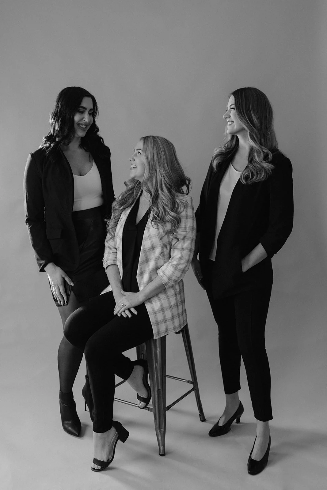 Three women in business attire smiling and conversing in a black-and-white studio setting, one seated on a stool and two standing.