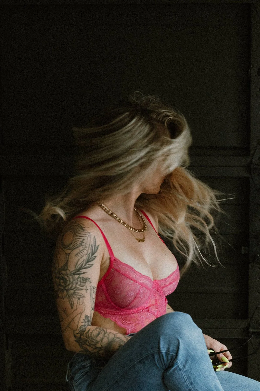 A woman with long blonde hair, tattoos on her left arm, wearing a pink lace bralette, gold chain necklace, and blue jeans, sitting against a dark background.
