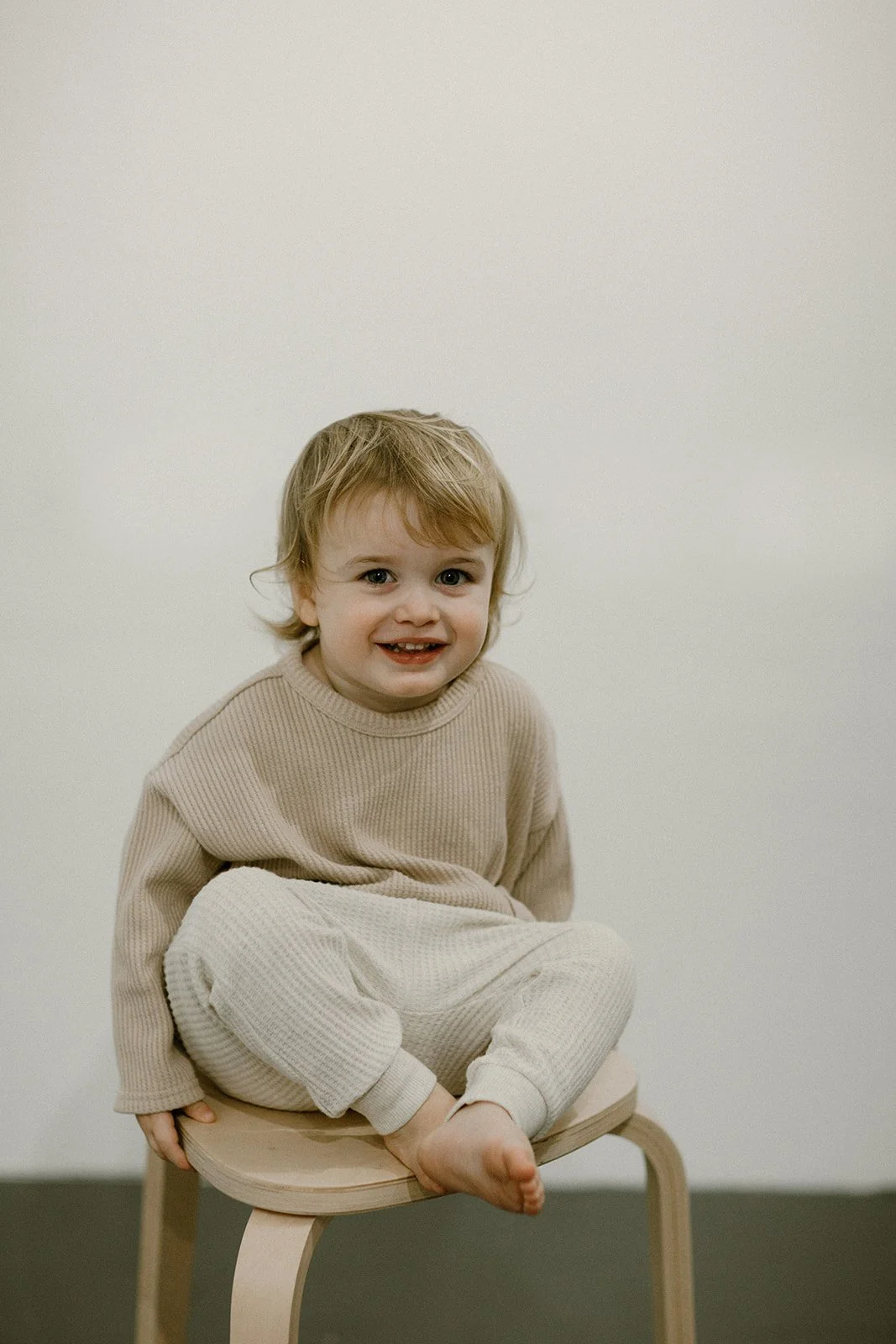 A young child sitting cross-legged on a wooden stool, smiling, wearing a beige ribbed sweater and matching pants, with a neutral plain background.