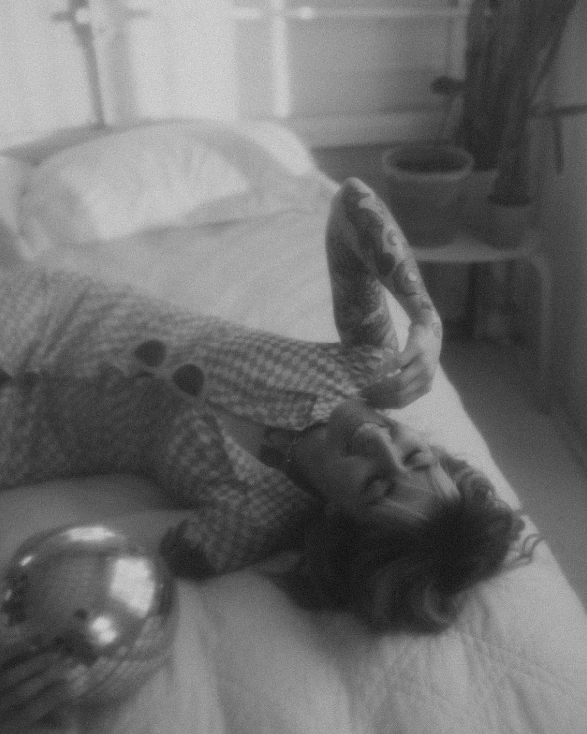 A person lying on a bed, holding a smartphone, with a large decorative ball nearby, in a room with a nightstand and a window in the background.