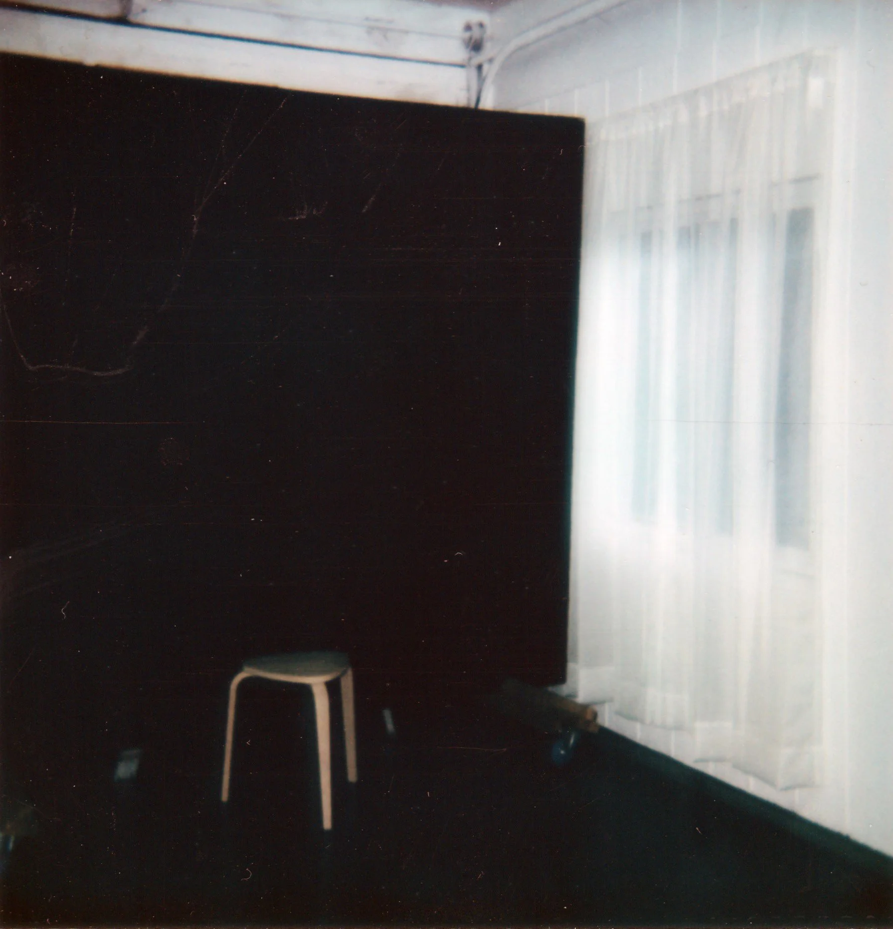 A photograph of a room with a black board, a white curtain, and a white stool on a dark floor.