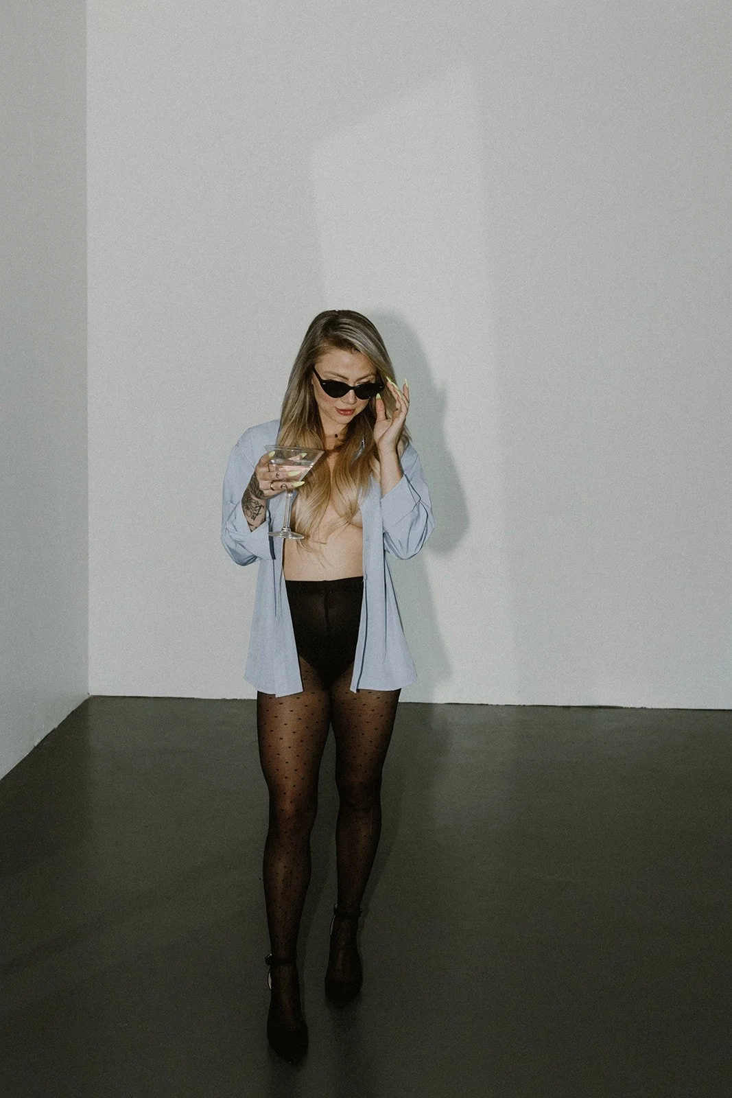 A woman with long blonde hair wearing sunglasses, an unbuttoned light blue shirt, black polka dot tights, and high heels, holding a martini glass and adjusting her sunglasses.