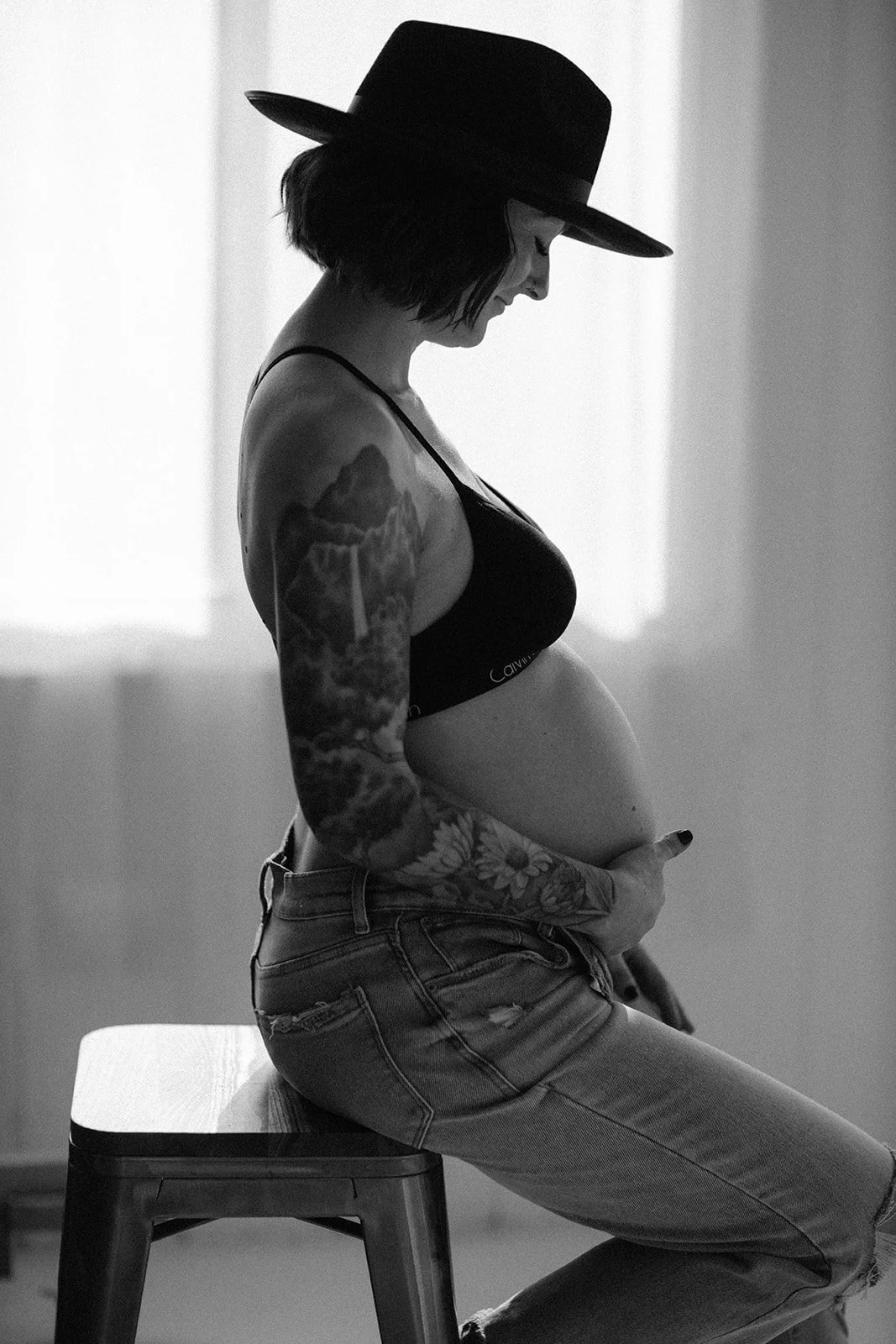 A woman wearing a hat, crop top, and ripped jeans sitting on a stool in profile, showcasing a tattooed arm and pregnant belly.