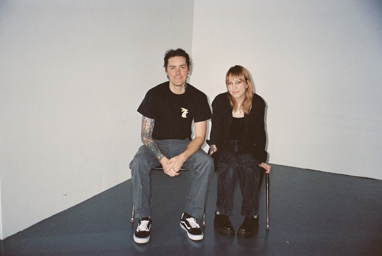 A man and a woman sitting on chairs against a white and light gray wall, both wearing black outfits with the woman wearing black platform shoes and the man in black sneakers. The man has tattoos on his left arm and short dark hair. The woman has red 