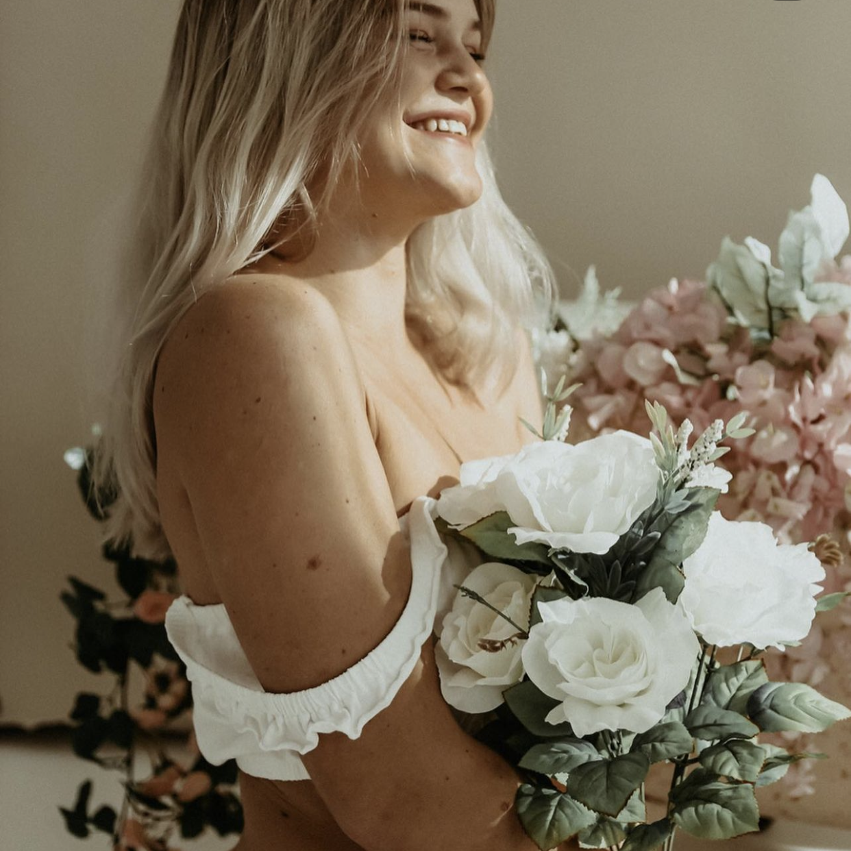 A woman with blonde hair smiling and holding a bouquet of white flowers, standing in front of a background of pink and white flowers.