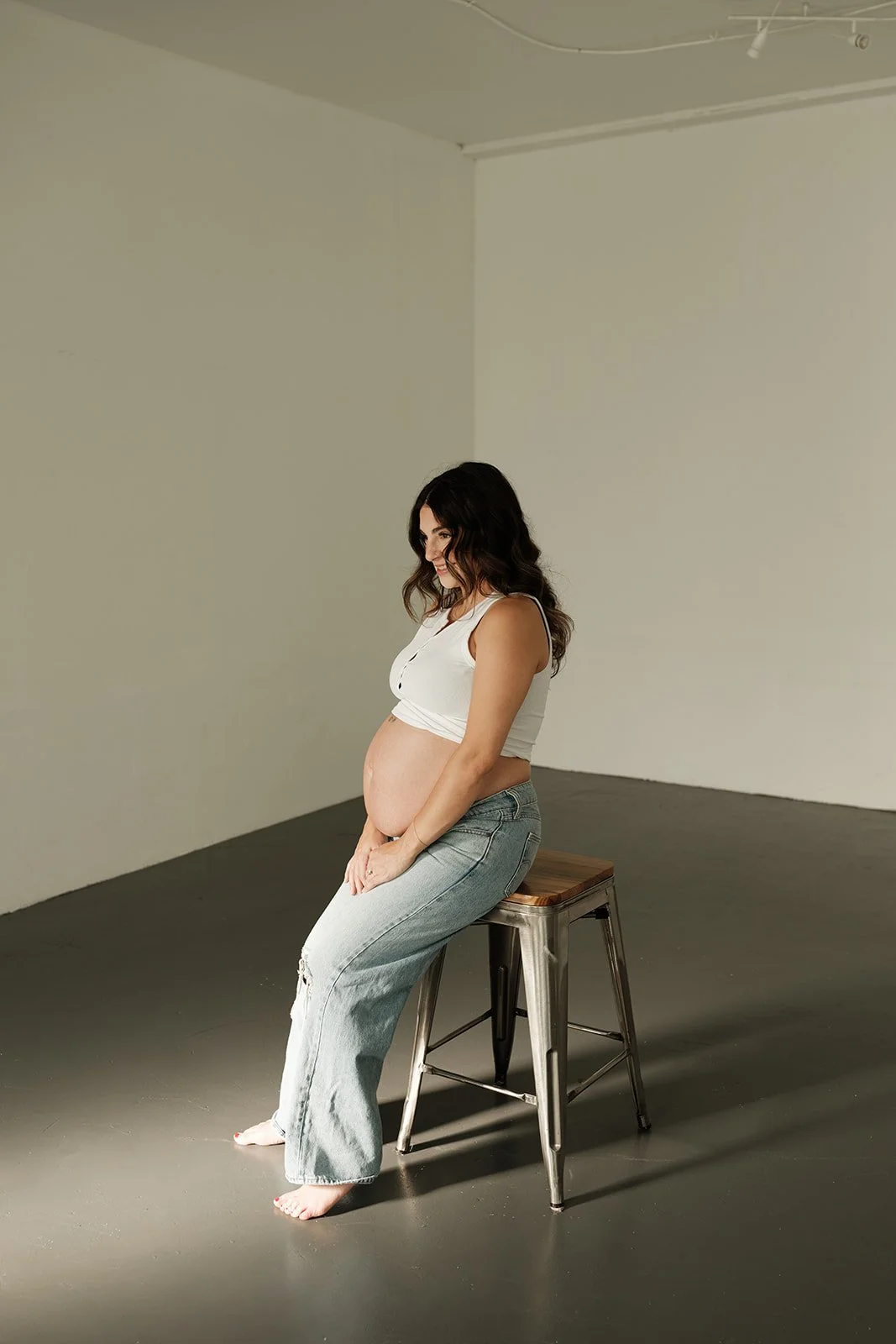 A pregnant woman sits on a metal stool in a minimalist studio with plain walls and dark flooring, looking down at her baby bump.