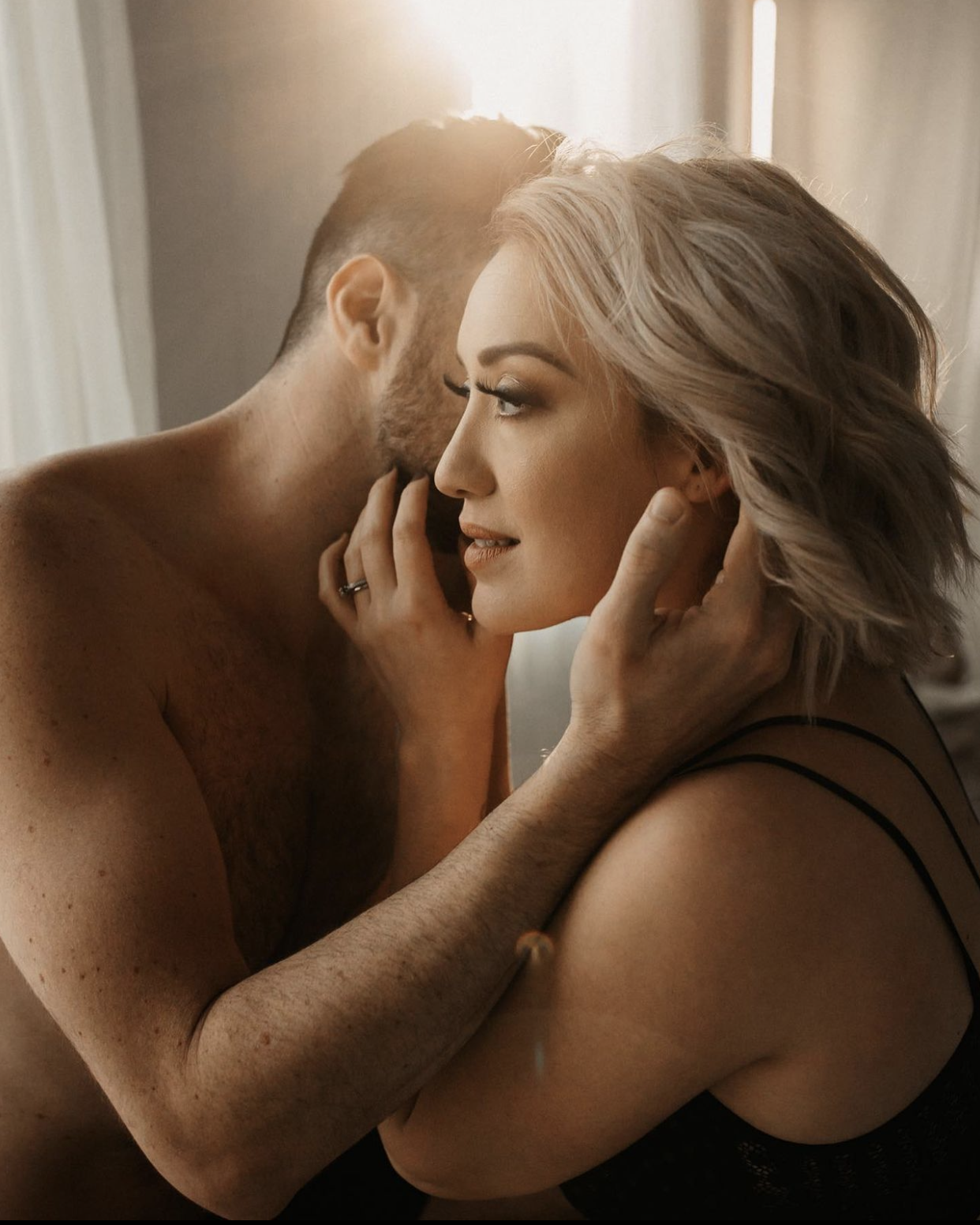 A shirtless man and a woman with blonde hair hold each other's faces close in an intimate moment indoors.