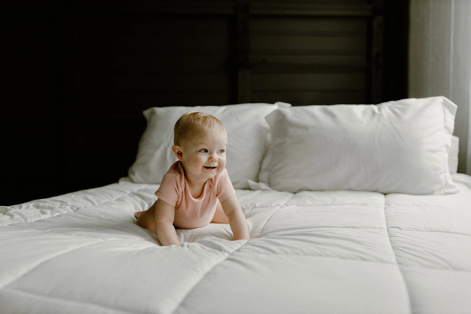 A smiling toddler crawling on a white bed with pillows, in a room with dark wooden walls and a sheer curtain on the right.