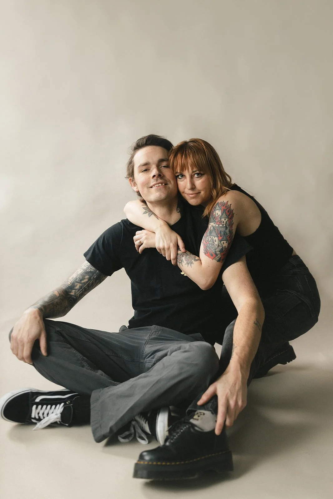 A couple with tattoos sitting on the floor, embracing each other, against a plain light-colored background.