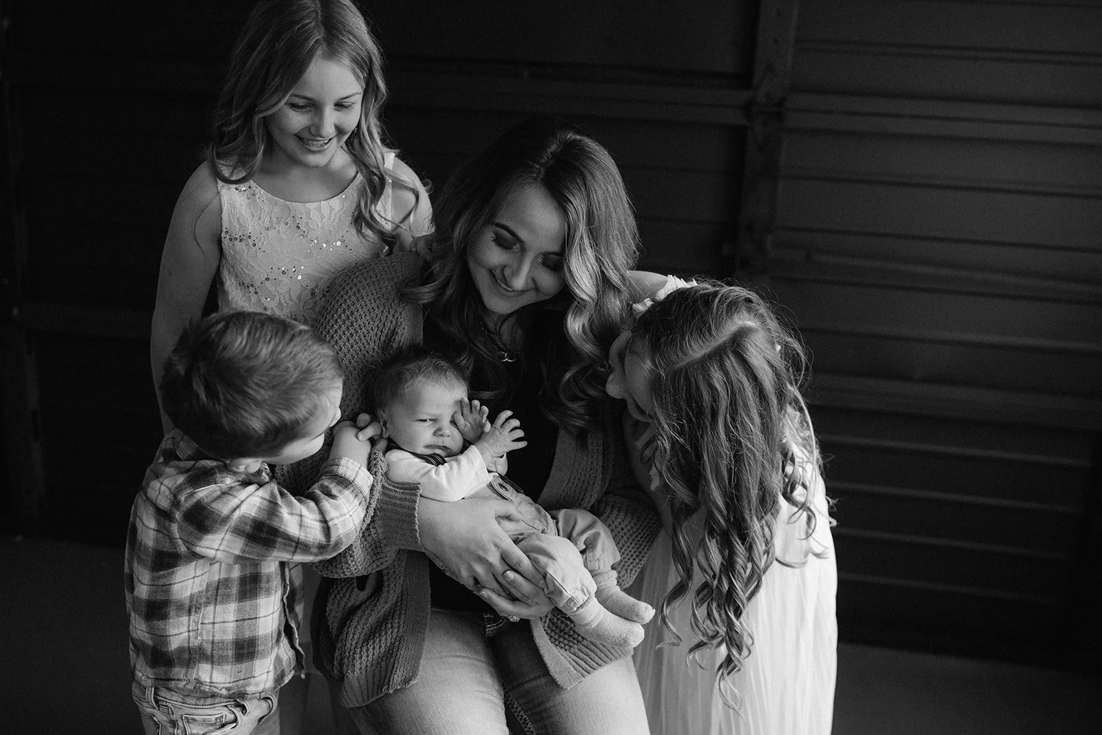 A woman holding a baby surrounded by four children, all smiling, in a cozy indoor setting.