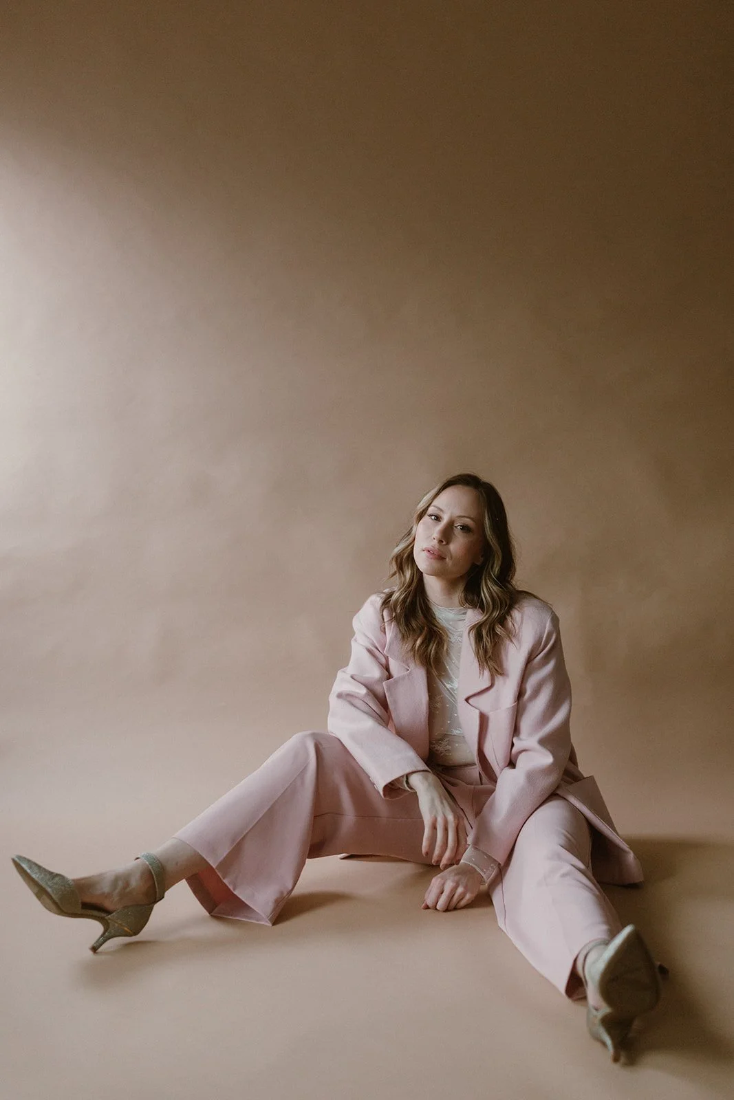 A woman in a light pink suit sitting on the floor against a plain beige background, with one leg extended and the other bent, wearing heels and looking at the camera.