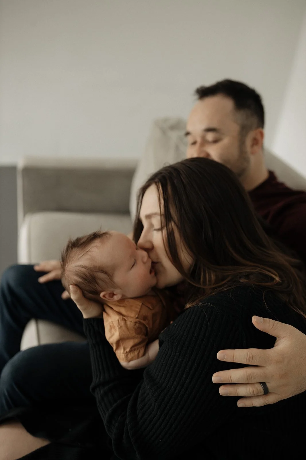 A woman and a man are sitting on a bed, sharing a tender moment as they kiss their newborn baby. The woman cradles the baby close to her face, and the man rests behind them, looking peaceful.