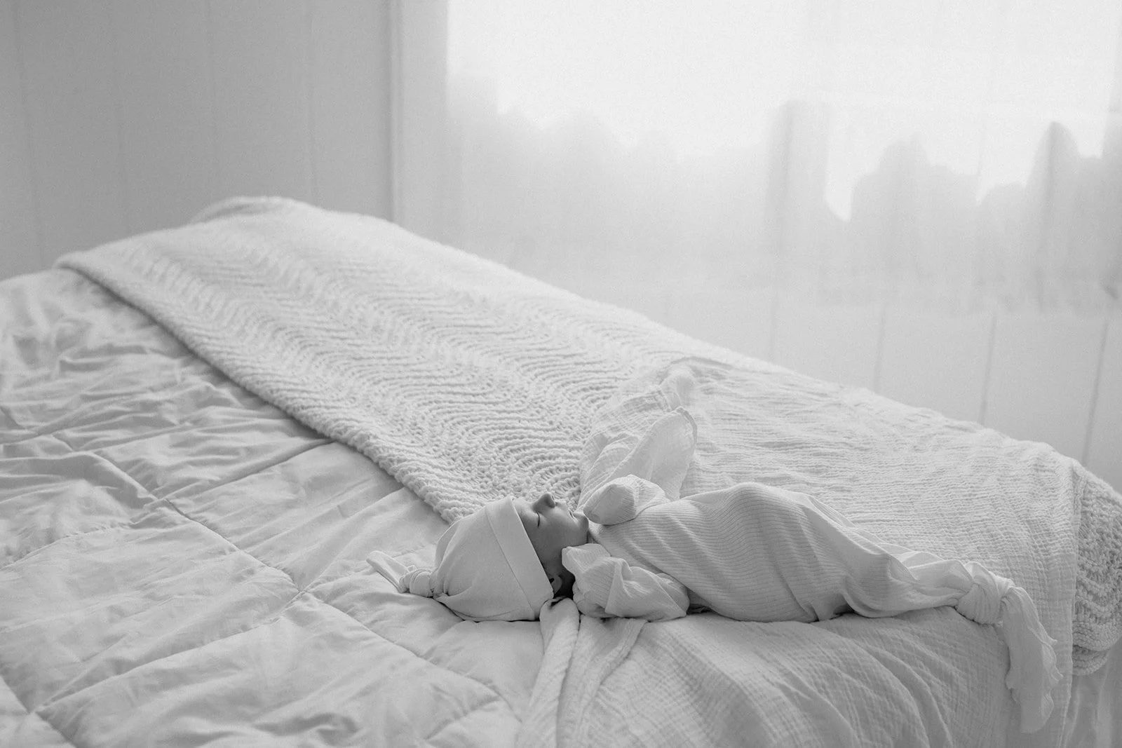 A newborn baby wrapped in a white blanket lies on a bed with white bedding, wearing a white hat and swaddling clothes, in a softly lit room.