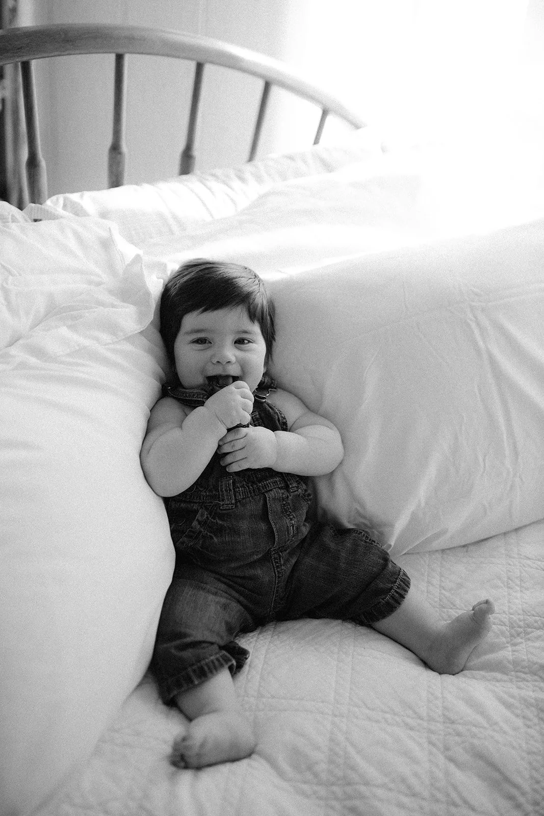 A young child sitting on a bed, smiling and holding their hand near their mouth, with pillows behind them.