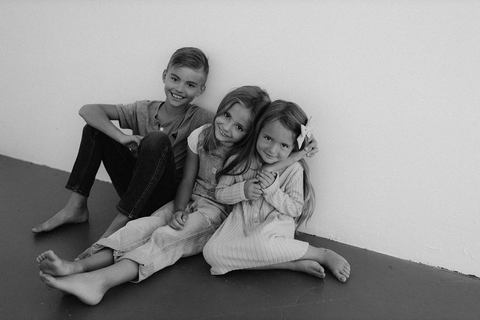 Three children, two girls and one boy, sitting on the floor and smiling at the camera, with plain wall background.