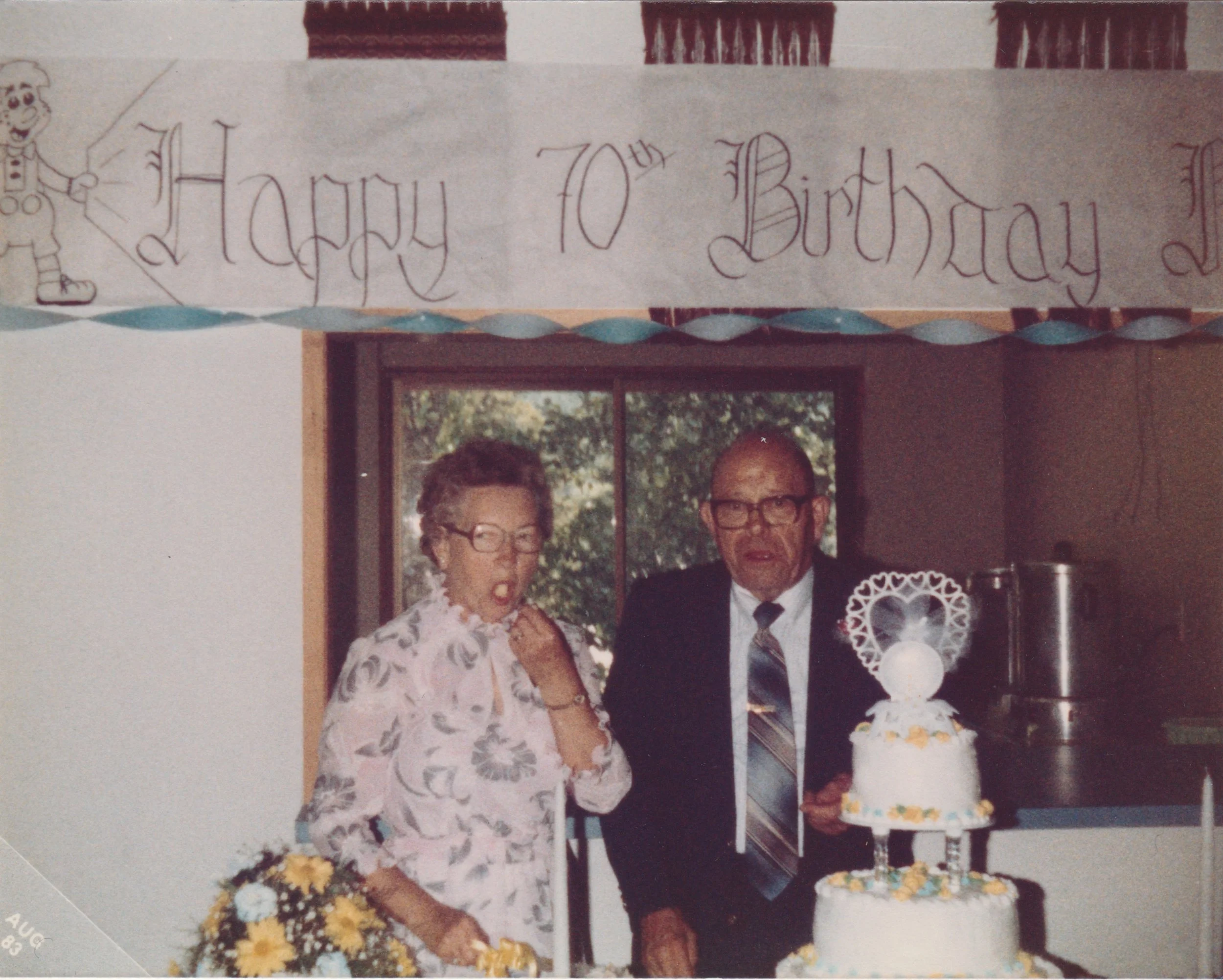 A birthday celebration with an elderly woman and man standing behind a decorated cake, with a banner above that says 'Happy 70th Birthday,' and the woman holding a bouquet of flowers.