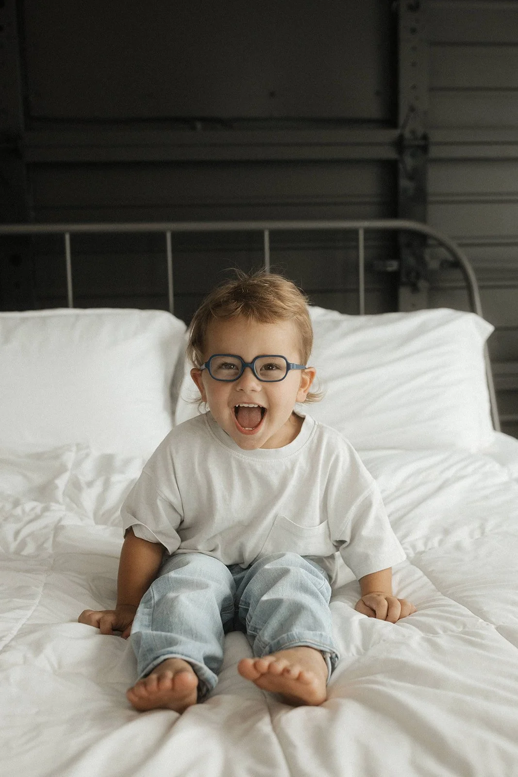 A young boy with glasses, smiling and sitting on a white bed with a dark metal headboard, in a room with dark paneled walls.