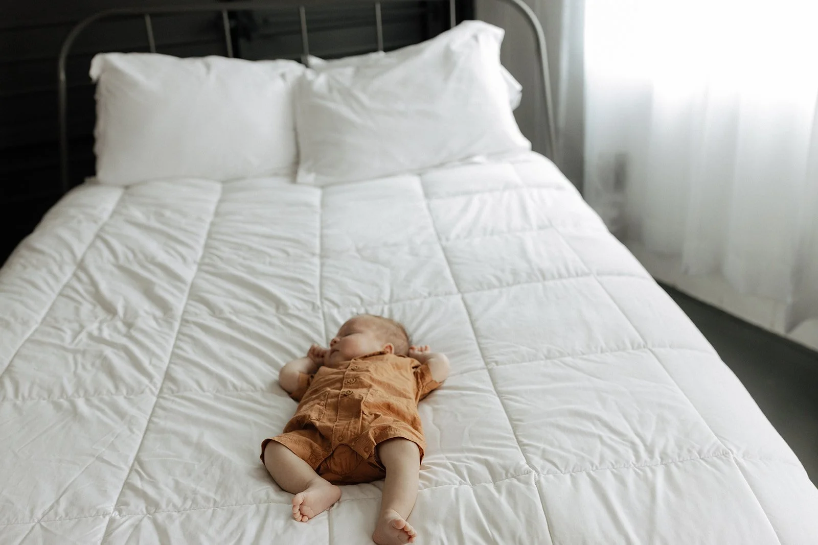 A baby in a brown outfit sleeping on a white bed with pillows and a headboard, near a window with curtains.