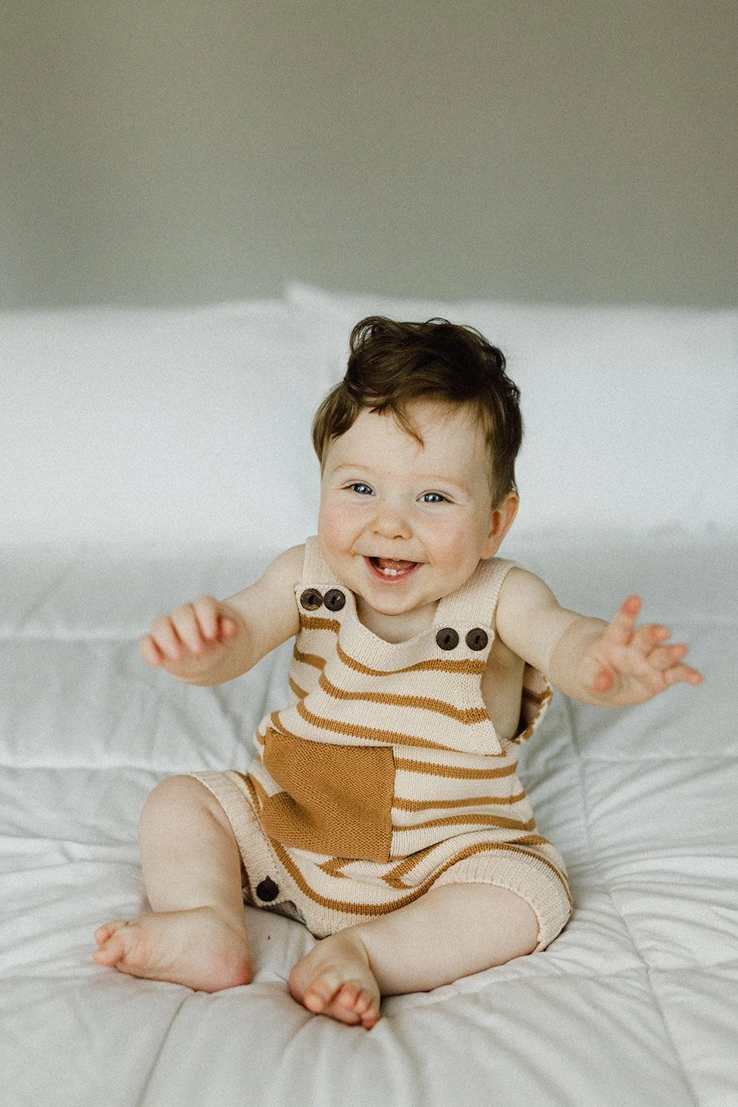 A smiling baby with curly hair sitting on a white bed, wearing beige and brown striped overalls.