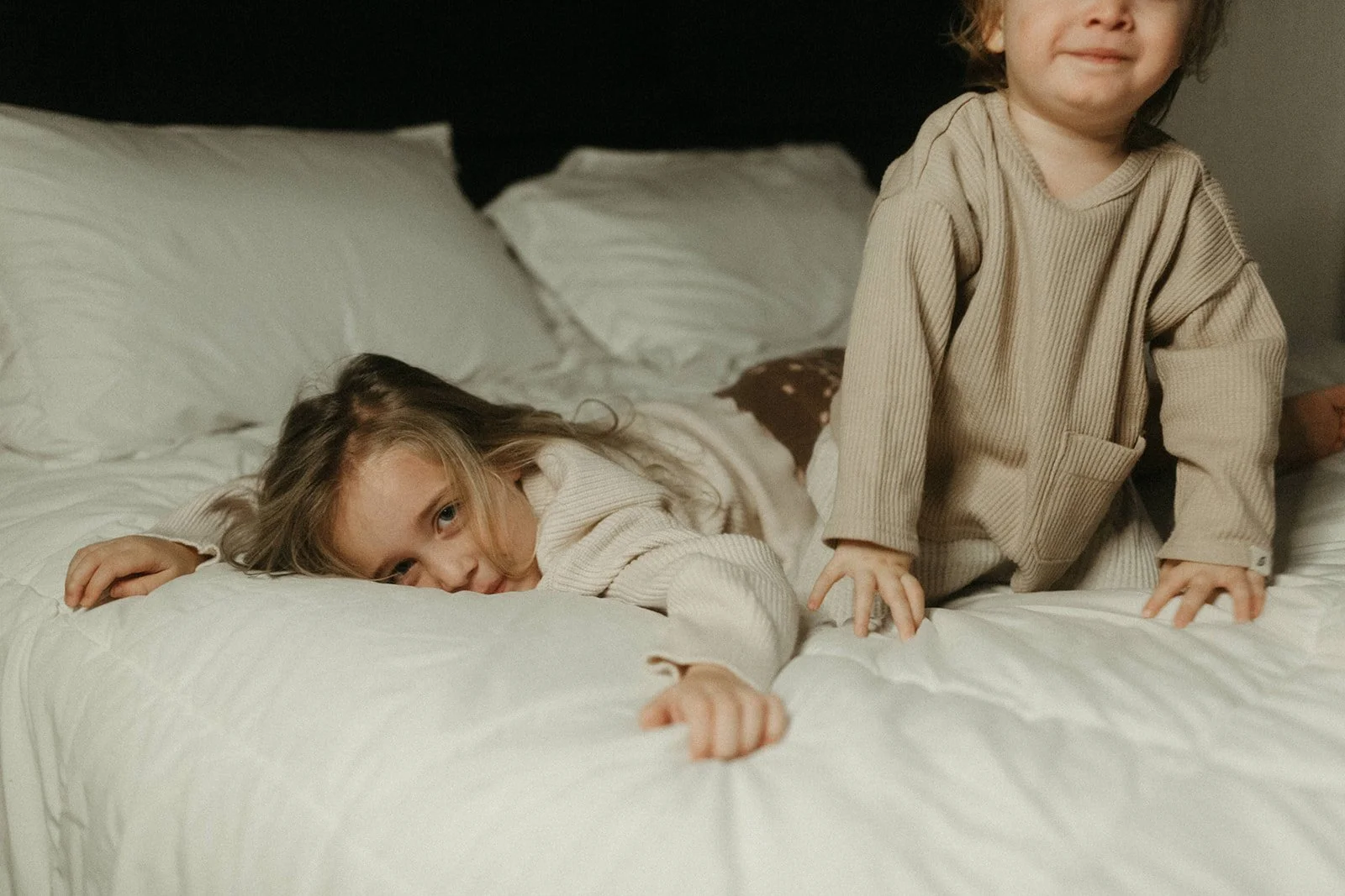 Two young children in beige pajamas on a bed with white bedding, one lying down resting her head on her arm, and the other kneeling.