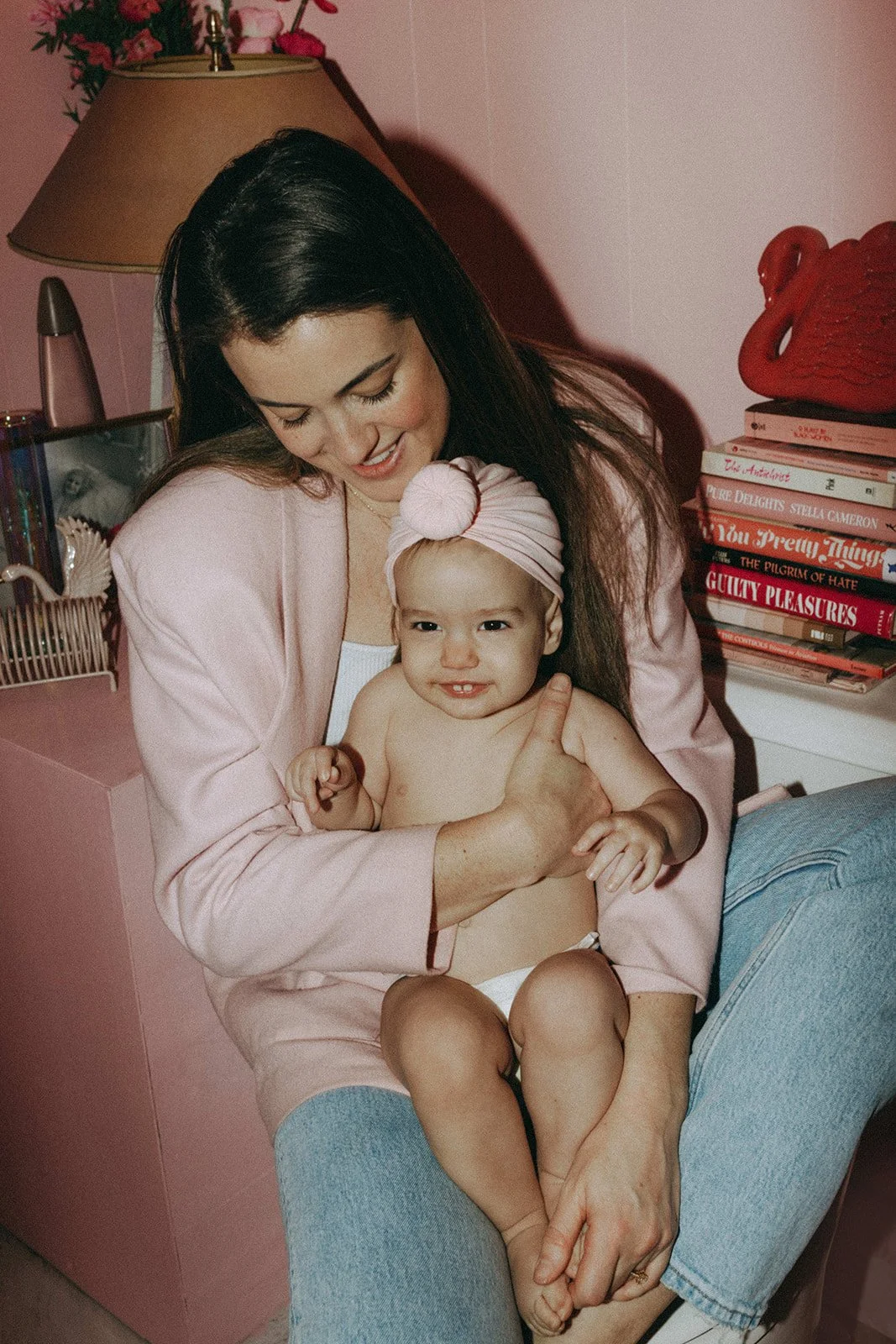 A woman holding a smiling toddler girl with a pink headband, in a pink-themed room with a lamp, books, and decorative items.