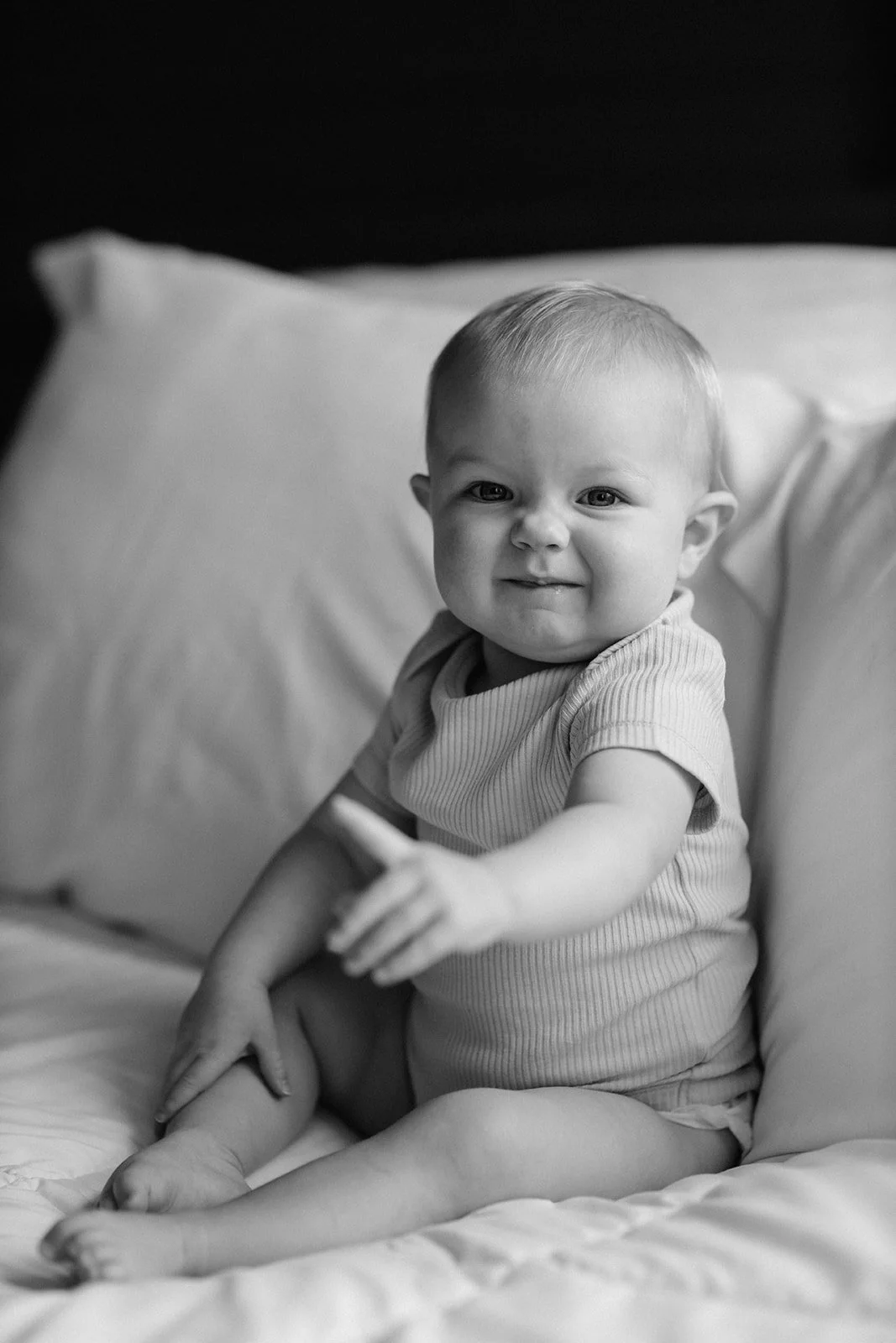 Black and white photo of a smiling baby sitting on a bed, pointing forward with one hand, looking at the camera.