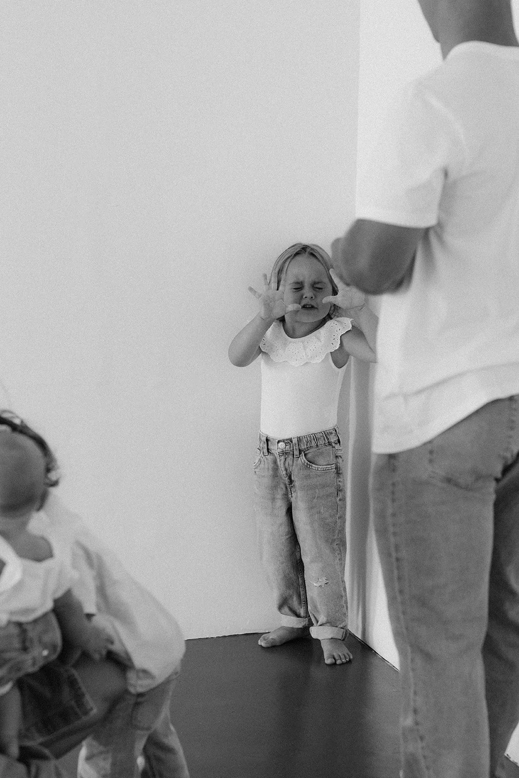 A young girl with a distressed expression, holding her hands near her face, leaning against a wall, as an adult stands nearby.