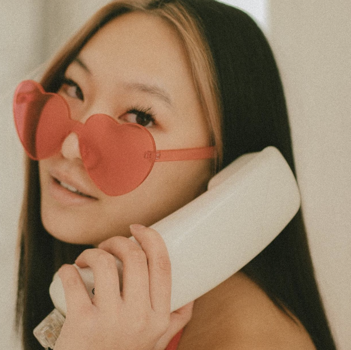 A young woman with long black hair wearing heart-shaped red glasses, holding a white vintage telephone receiver to her ear, smiling slightly.