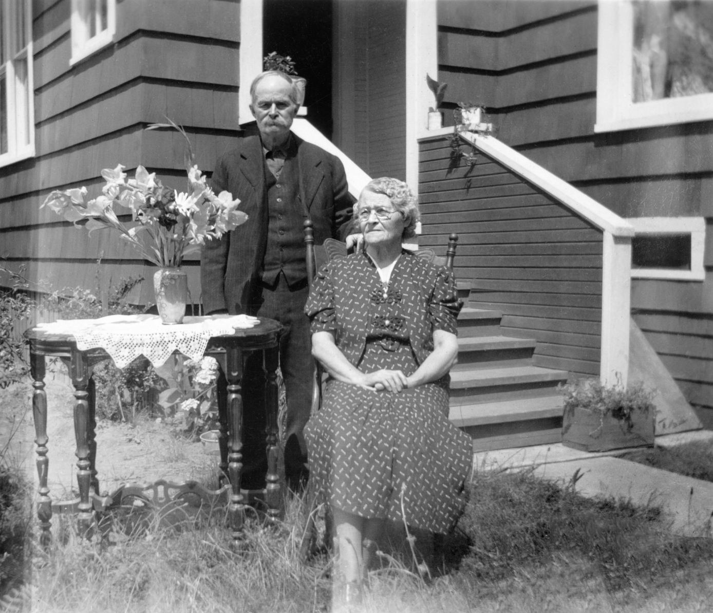 An elderly woman sitting on a wooden chair outdoors in front of a house, with a man standing behind her. The woman is wearing a patterned dress and glasses, and the man is dressed in a suit. A table with a vase of flowers and a doily is beside her. The house has siding and a staircase leading to the door.