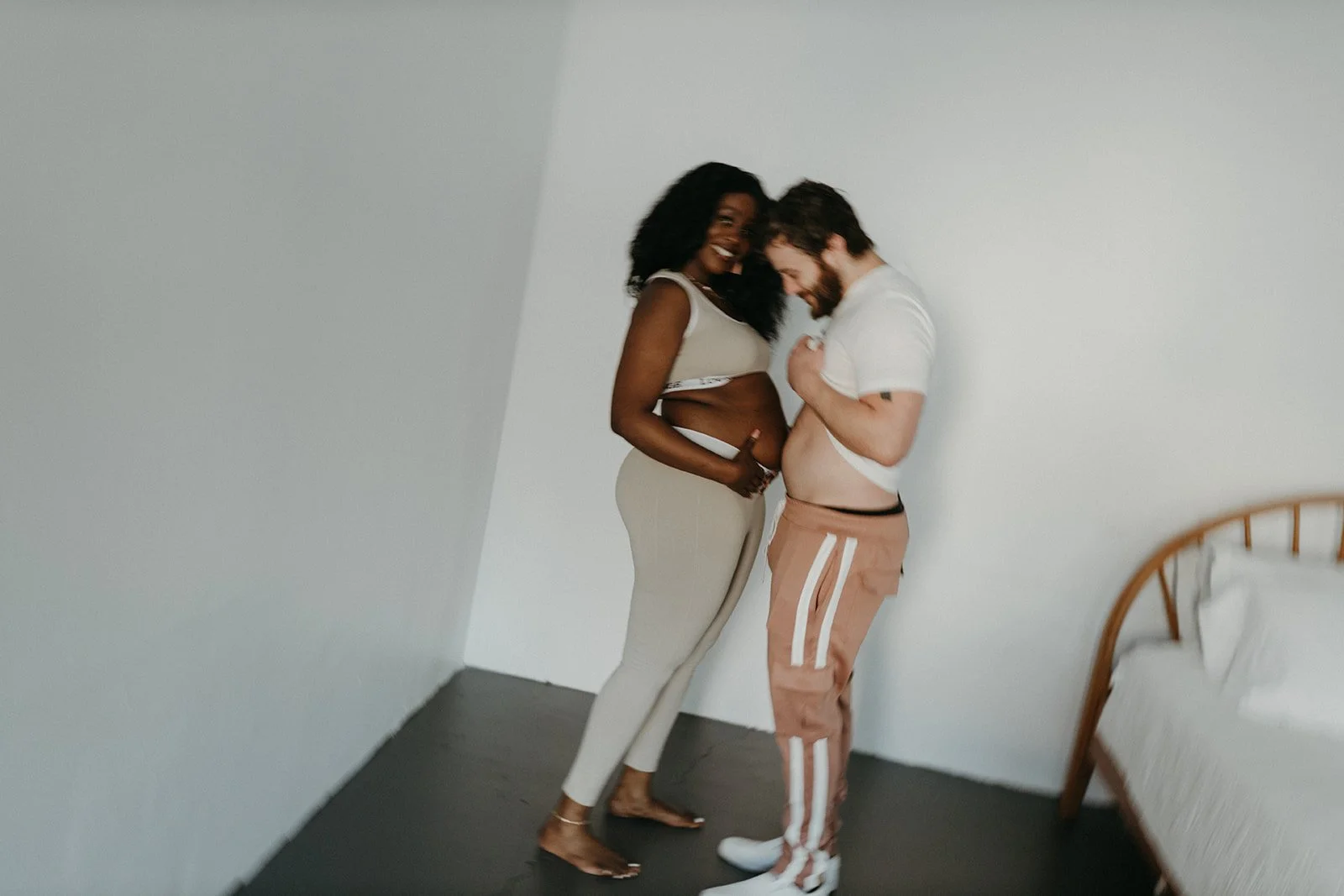 A happy couple, a woman with dark curly hair and a man with a beard, standing close together in a bedroom, smiling and touching their stomachs.