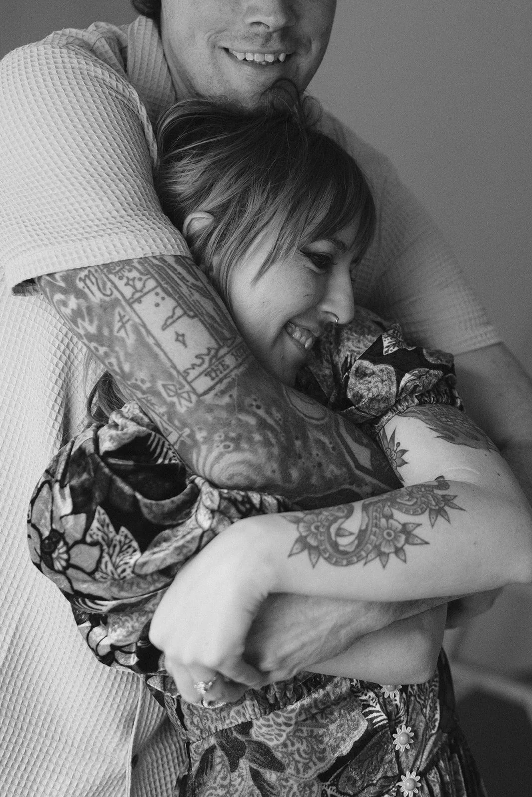 A black-and-white photo of a smiling person with tattoos hugging another person with short hair and tattoos, who is also smiling in a joyful embrace.