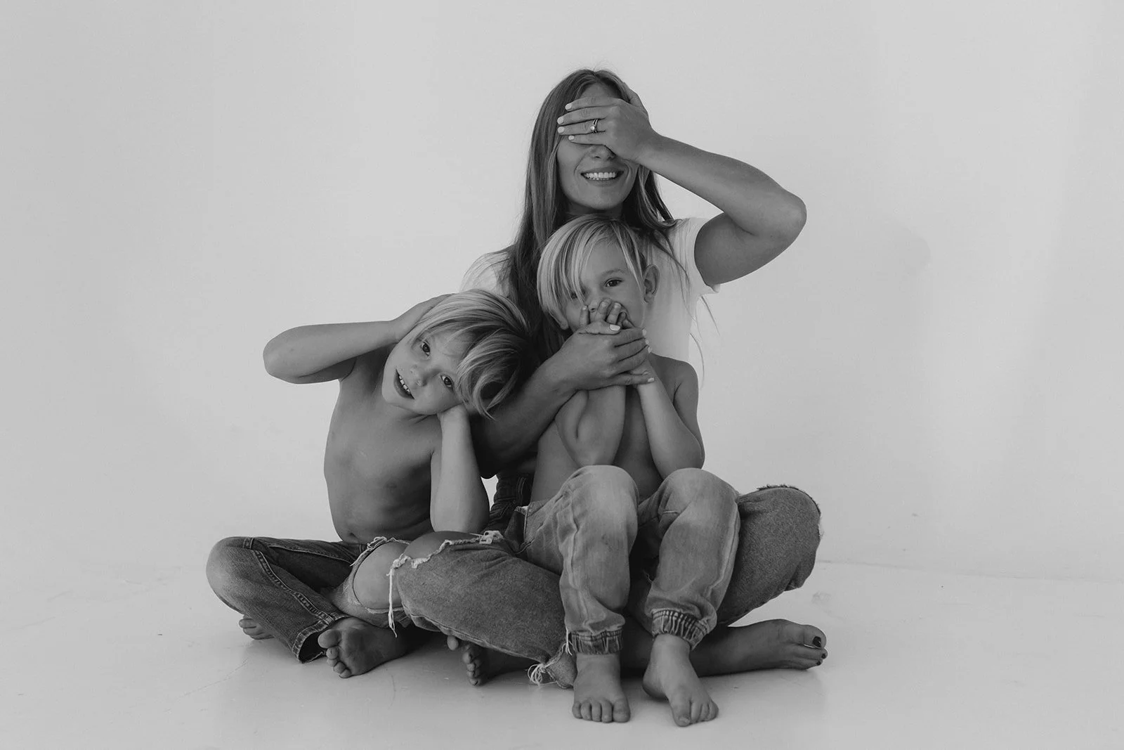 Black and white photo of a woman with two shirtless children, all sitting on the floor against a plain wall. The woman covers her eyes with her right hand, smiling, while the children, one leaning on her and the other in her lap, cover their mouths w