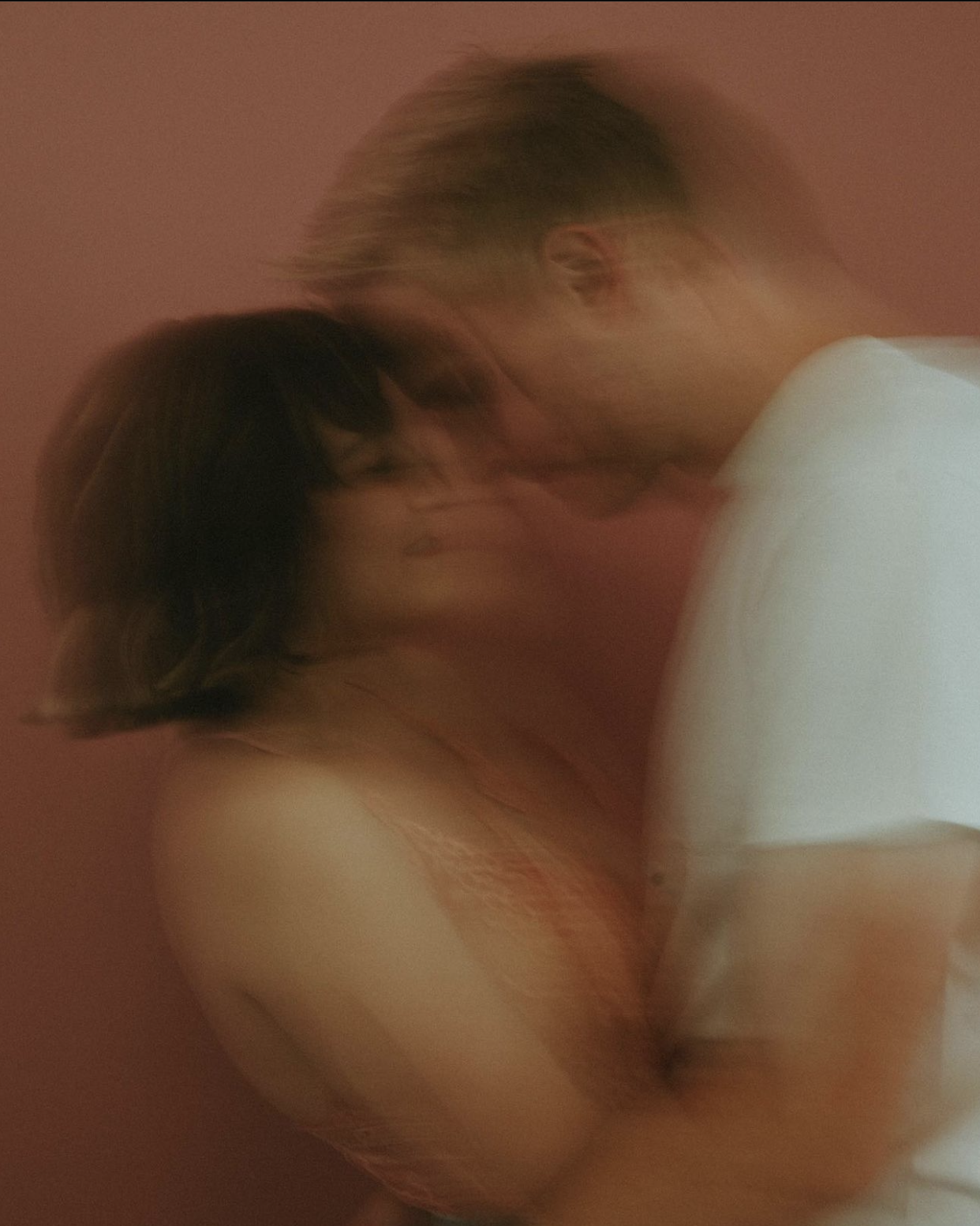 A blurry photo of a man and woman embracing each other close, with the man's face near the woman's face against a pink background.
