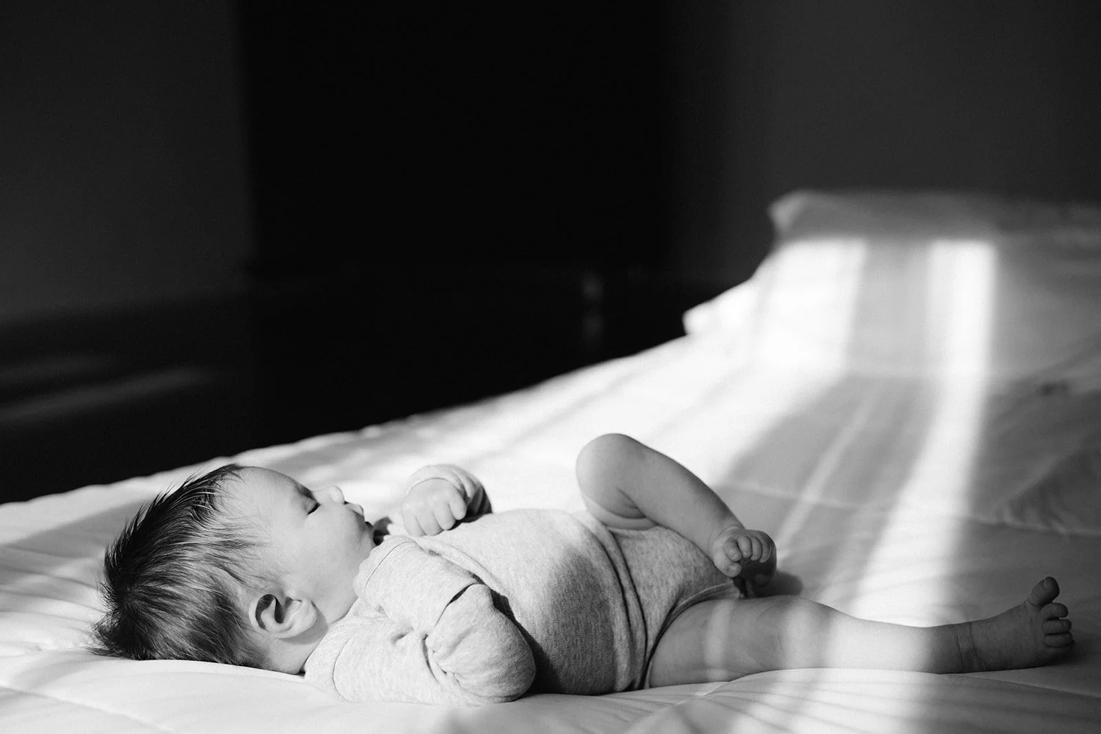 A baby lying on a bed, looking up with a hand near their face, in black and white.
